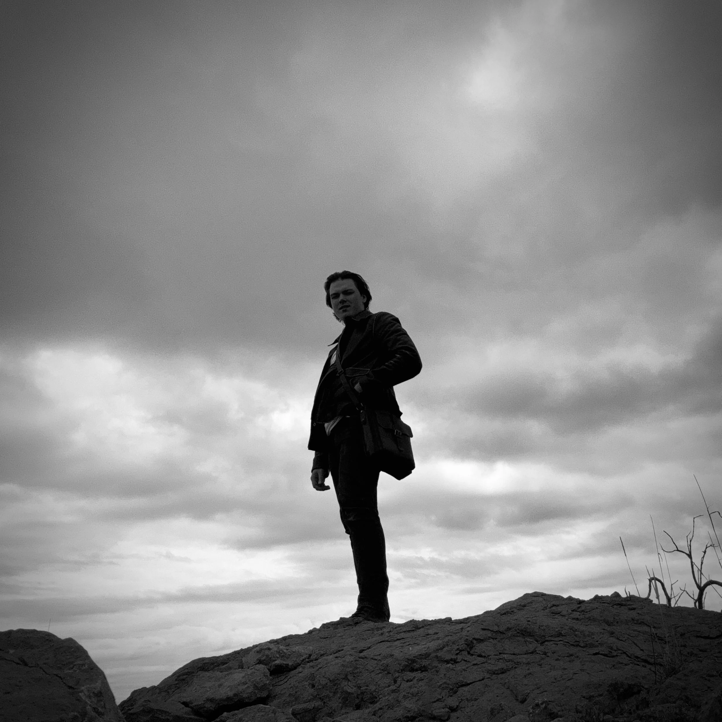 Black and white photo of a man standing on rocks outdoors under cloudy sky, with a serious expression, wearing dark clothing.