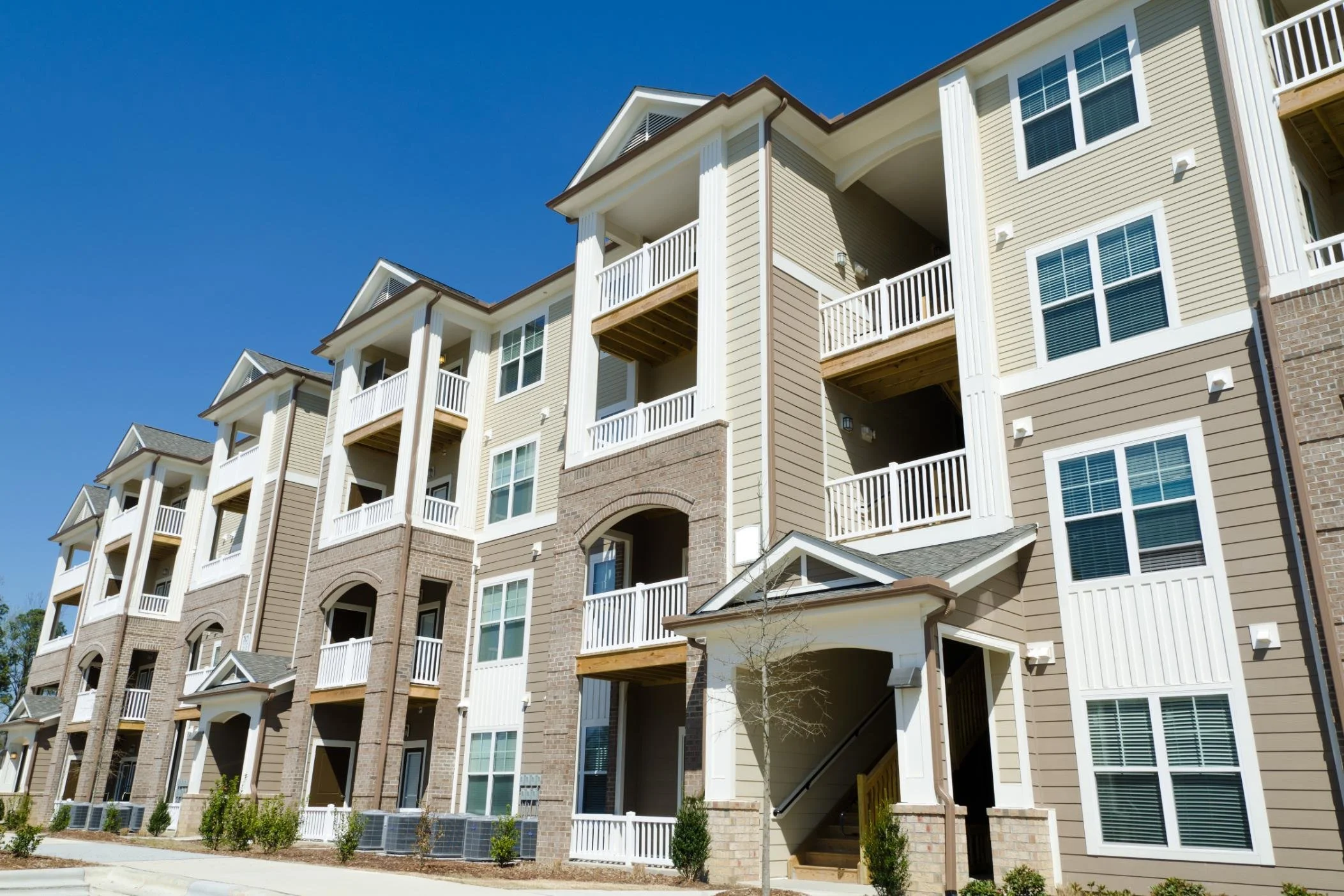 Multi-story residential apartment building with balconies, beige and brown exterior, and white trim under a clear blue sky.