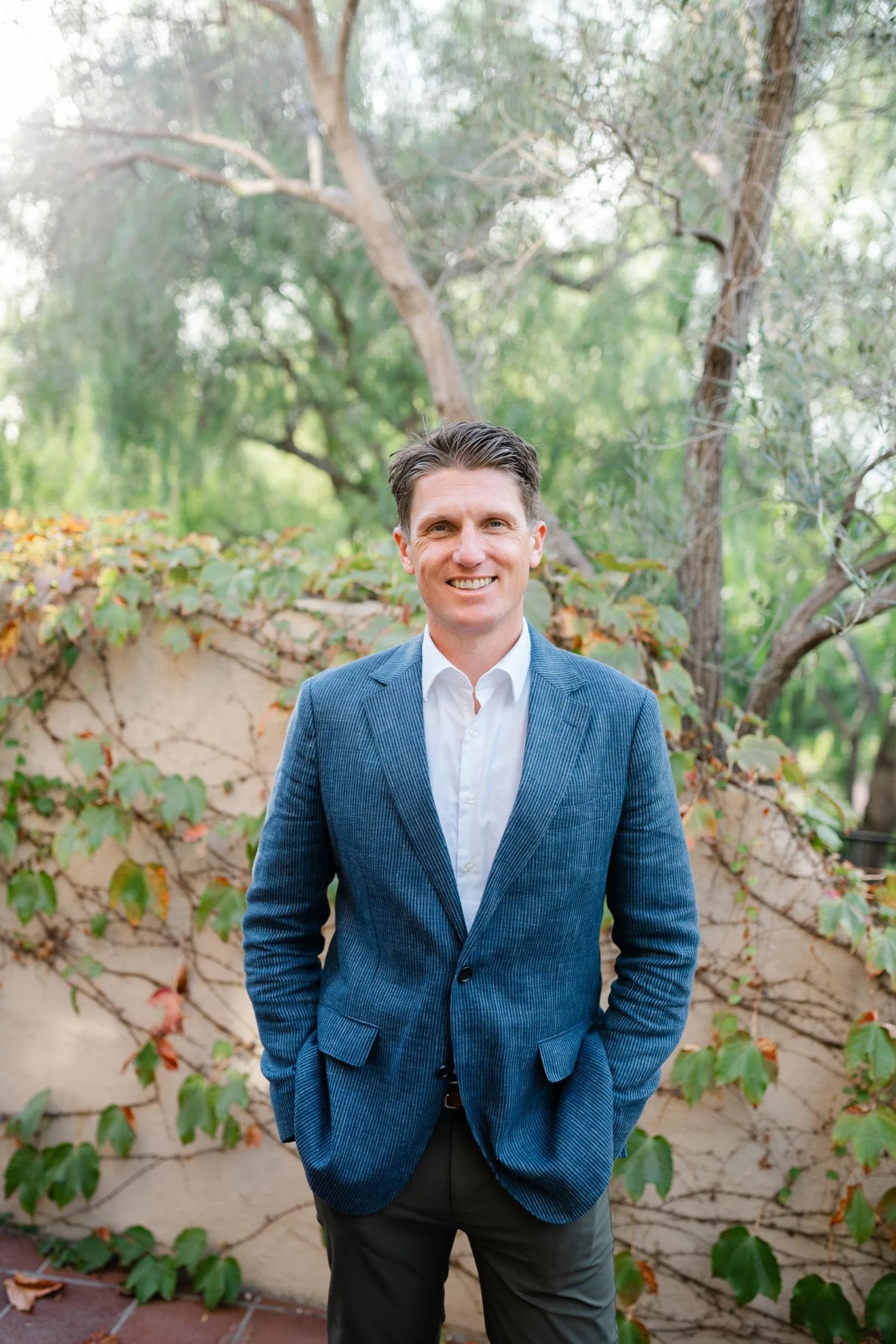 A man in a blue blazer and white shirt standing outdoors in front of a tree and a wall with climbing plants, smiling at the camera.