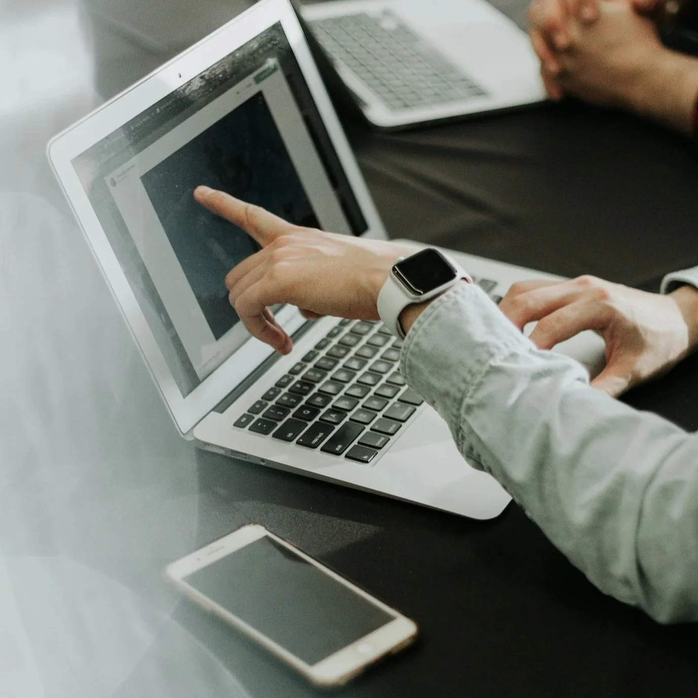 Person using a tablet with a smartwatch on their wrist, pointing at the screen, with a smartphone and another device on the table.
