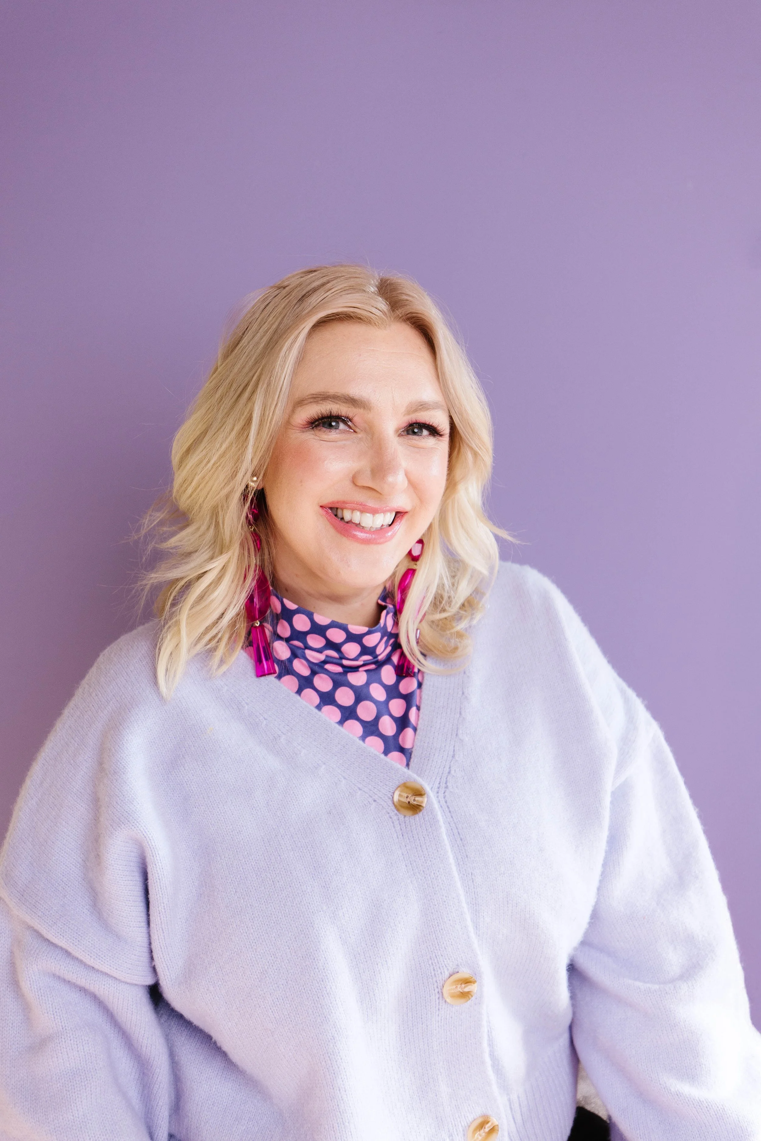 A woman with blonde hair wearing a white cardigan with large buttons, a pink polka dot top, and pink earrings, smiling against a purple background.