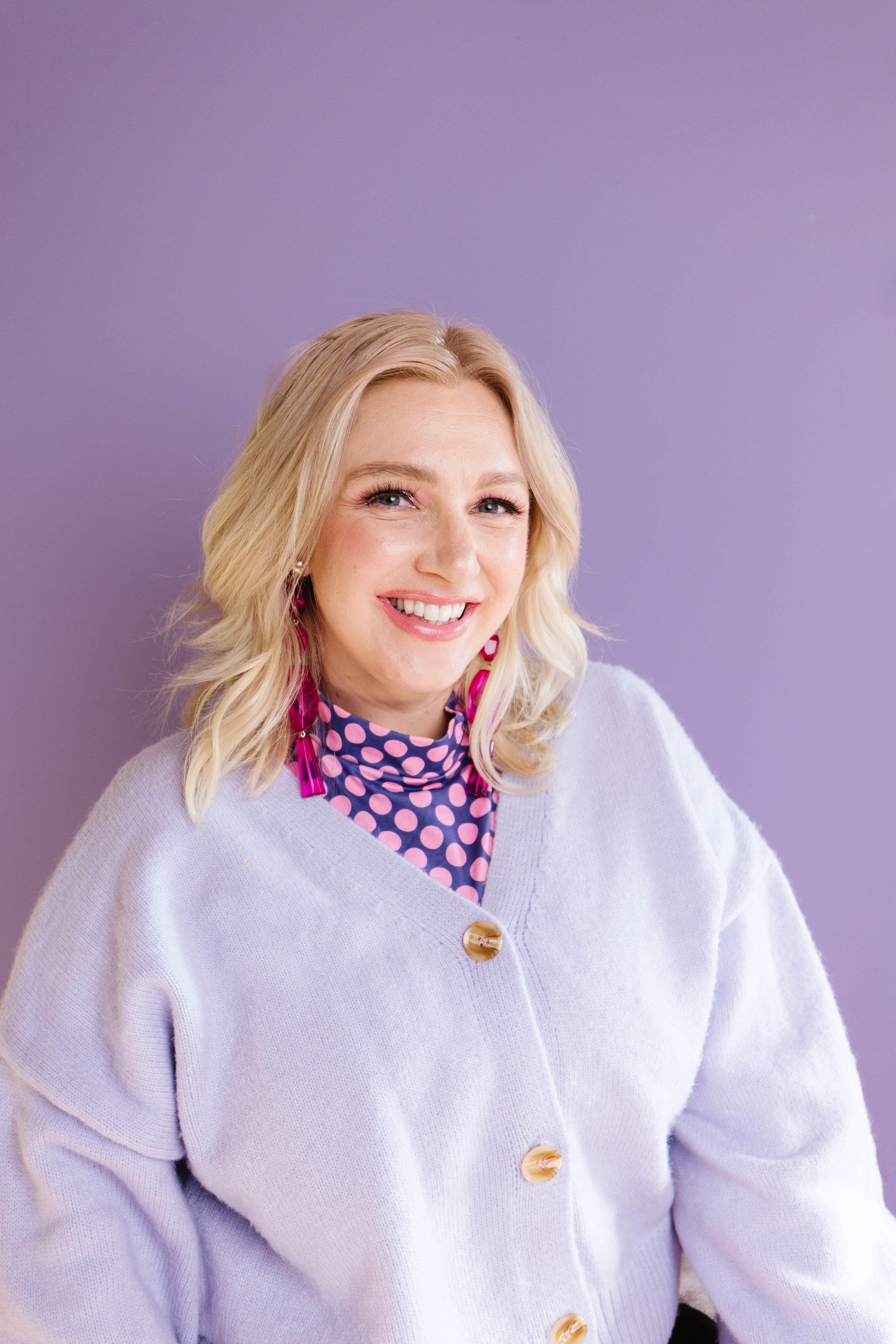 A woman with blonde hair smiling, wearing a light purple cardigan with large buttons, a pink polka dot top, and pink earrings, standing against a purple background.