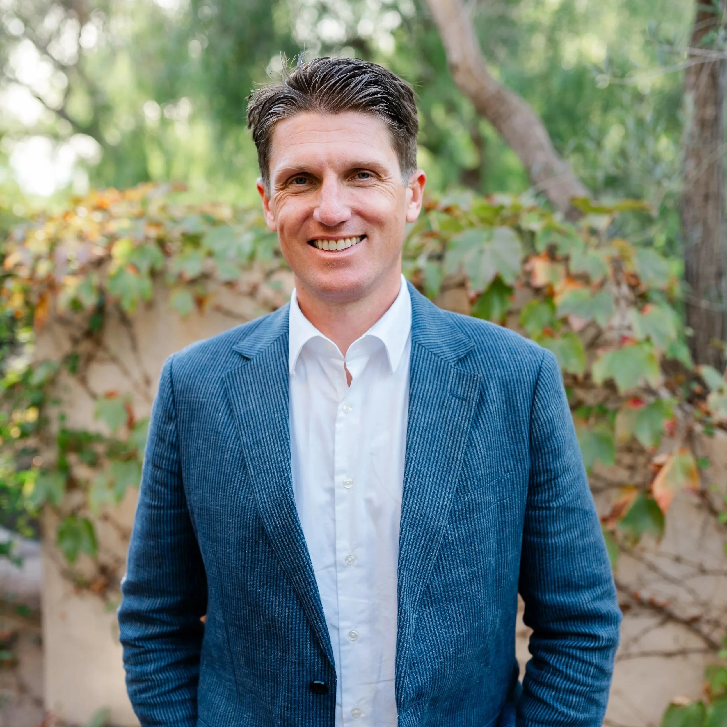 A man in a white shirt and blue blazer smiling outdoors with green foliage and trees in the background.