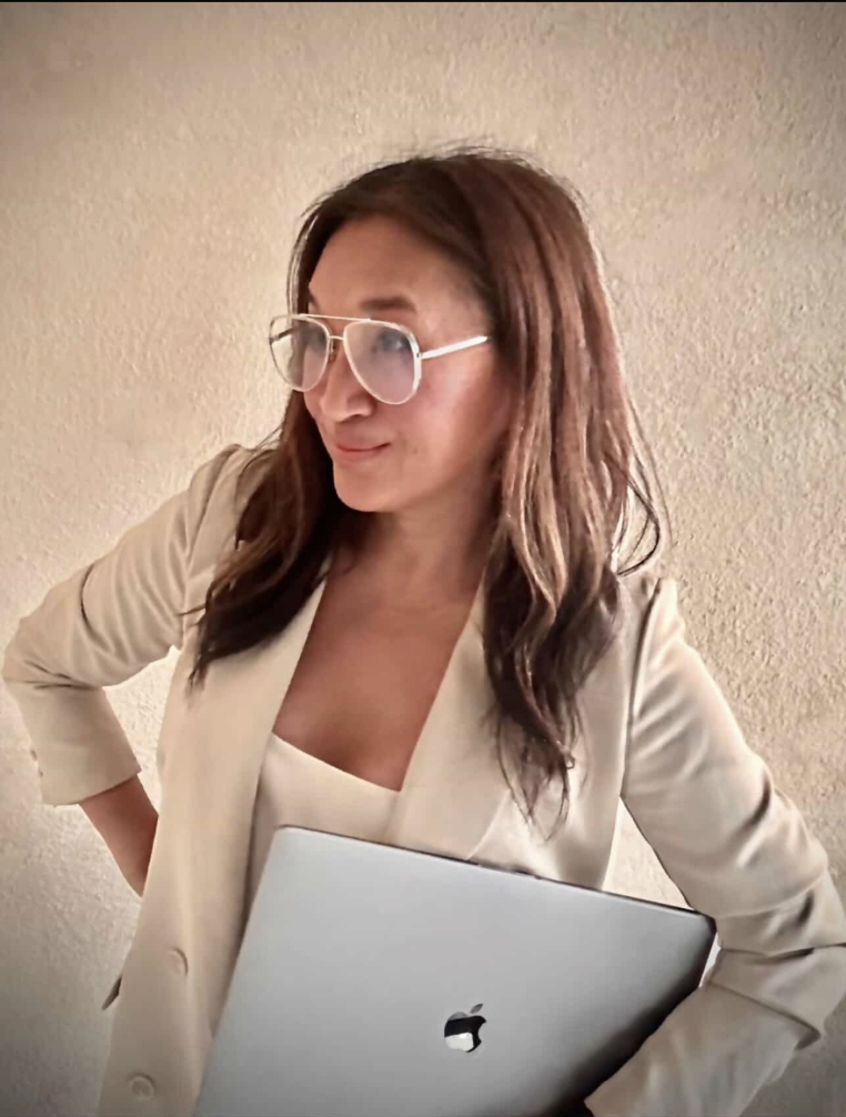 A woman with long brown hair, wearing glasses and a beige blazer, holding a silver Apple MacBook, standing against a textured beige wall.