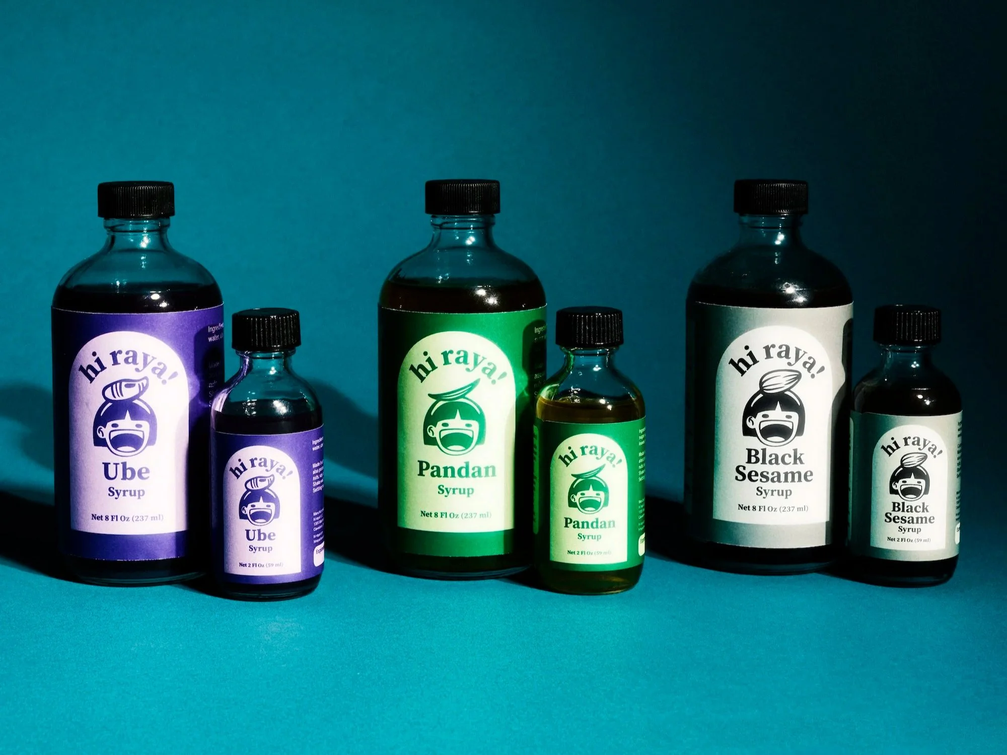 Bottles of syrup labeled 'hi raya!,' in flavors Ube, Pandan, and Black Sesame, arranged on a blue surface with a dark blue background.