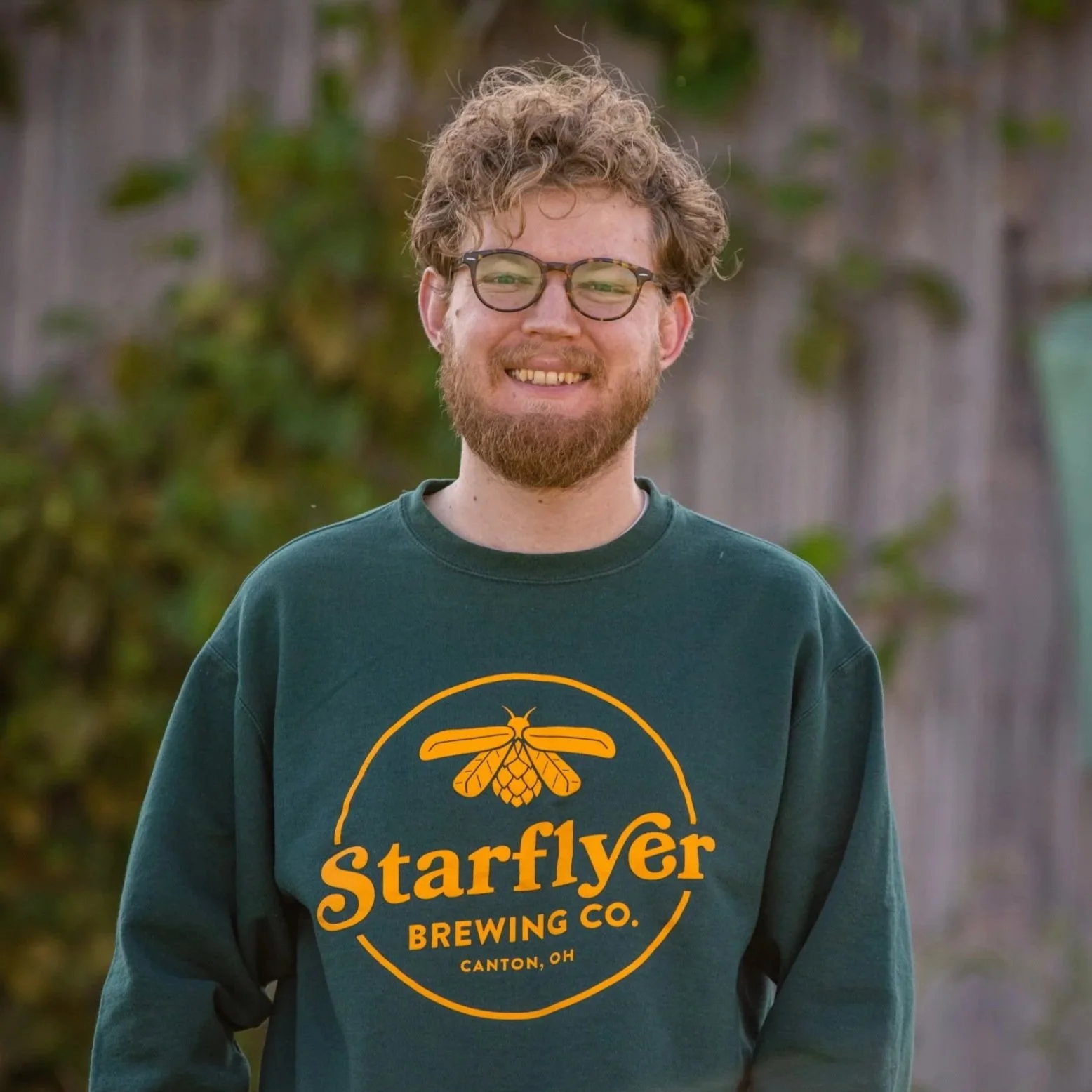 A smiling man with curly hair and glasses wearing a dark green sweatshirt with a Starflyer Brewing Co. logo, featuring a bee and hops, in Canton, Ohio.