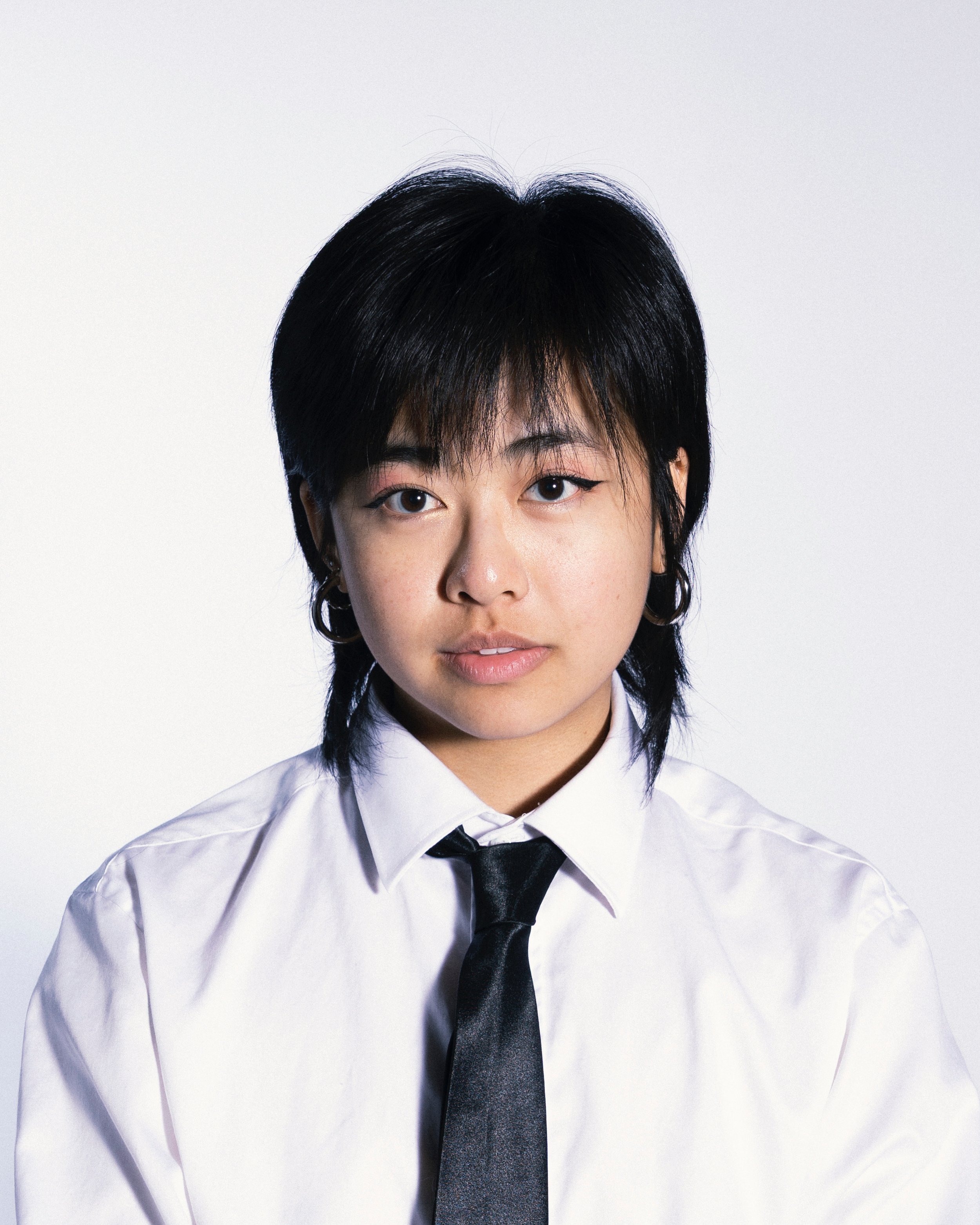 A young woman with short black hair, wearing a white shirt and black tie, against a plain light background.