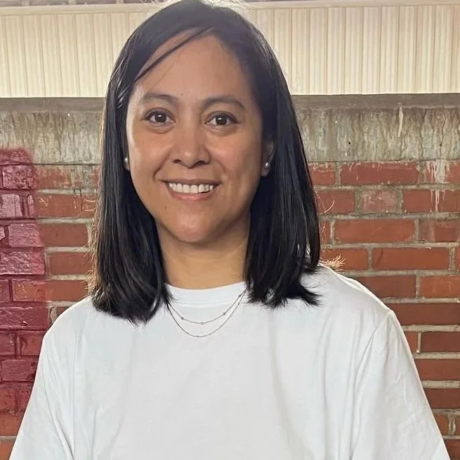 A woman with shoulder-length black hair wearing a white shirt and small earrings, smiling in front of a brick wall background.