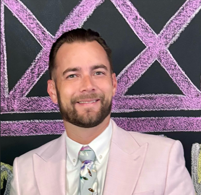 A smiling man with a beard and short hair, wearing a light pink suit jacket, a white shirt, and a patterned tie, standing in front of a dark background with large purple chalkboard drawings.