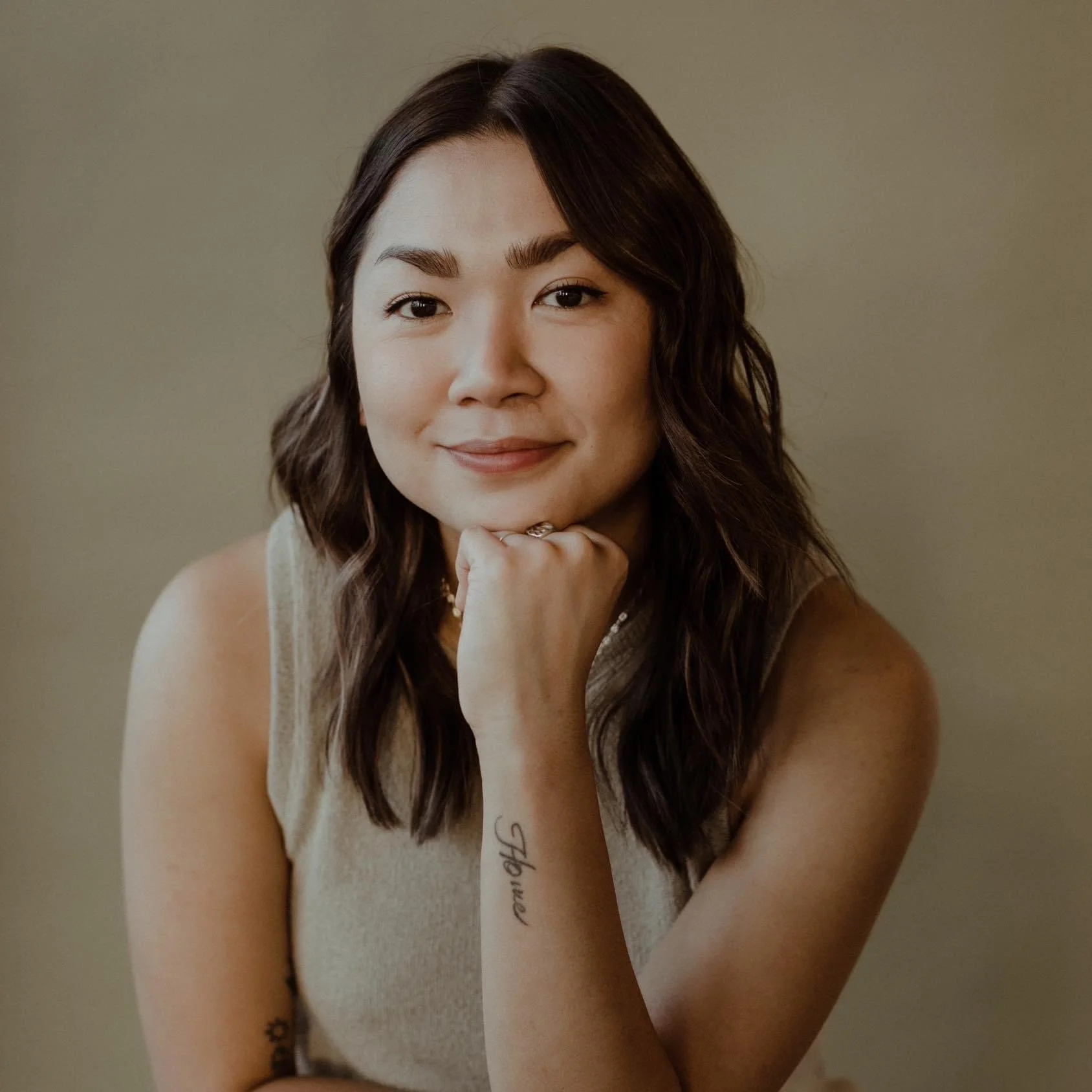 A woman with shoulder-length brown hair, sitting against a plain beige background, resting her chin on her hand, smiling softly.