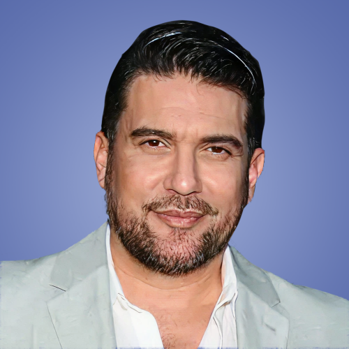 Close-up of a man with dark, slicked-back hair and a beard, wearing a light-colored blazer over a white shirt against a plain blue background.