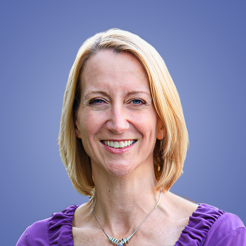A woman with shoulder-length blonde hair smiling at the camera, wearing a purple top with ruffled sleeves and a silver necklace, against a blue background.
