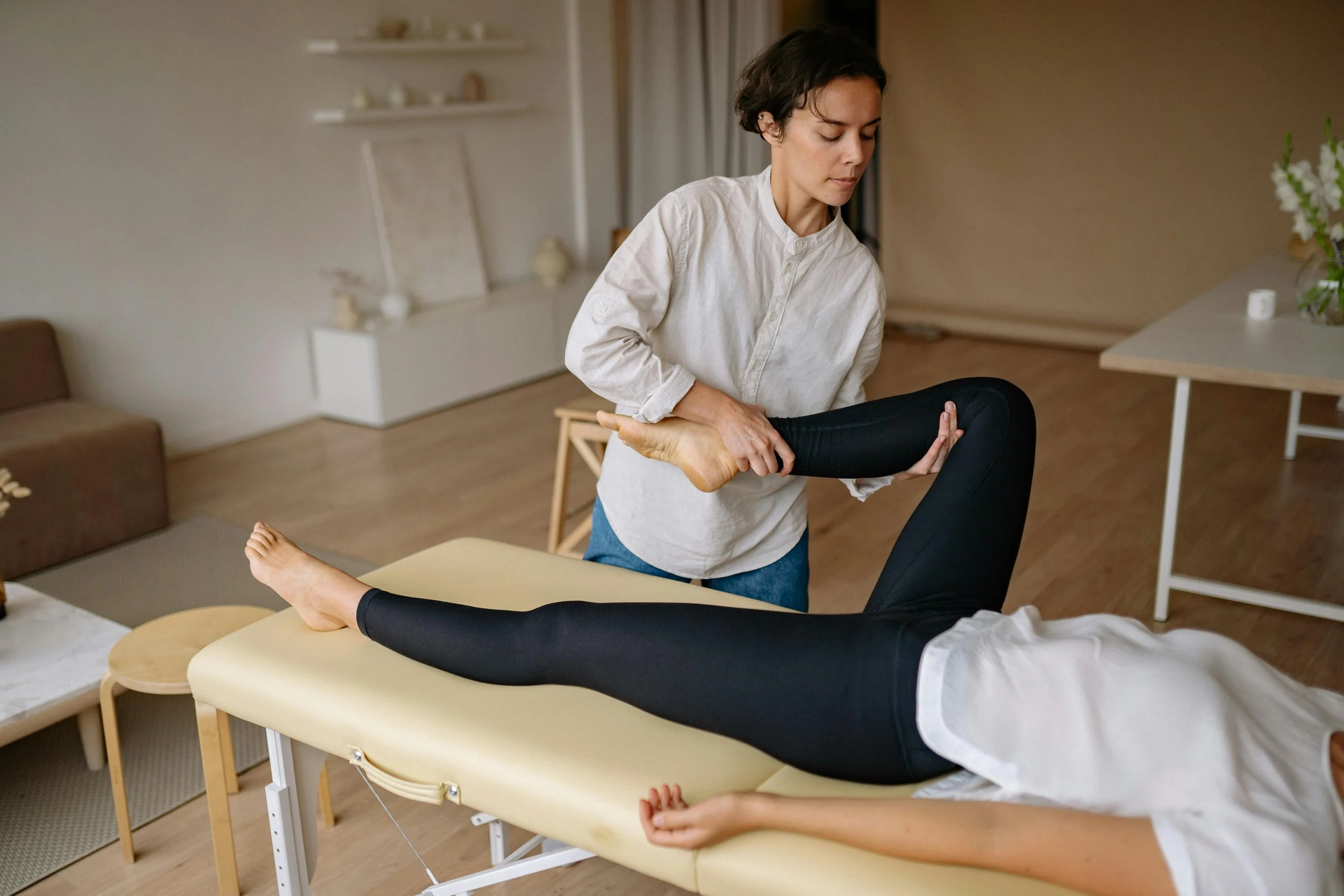 A woman receiving physical therapy while lying on a treatment table, holding her leg in a bent position as another woman assists her by holding her ankle.