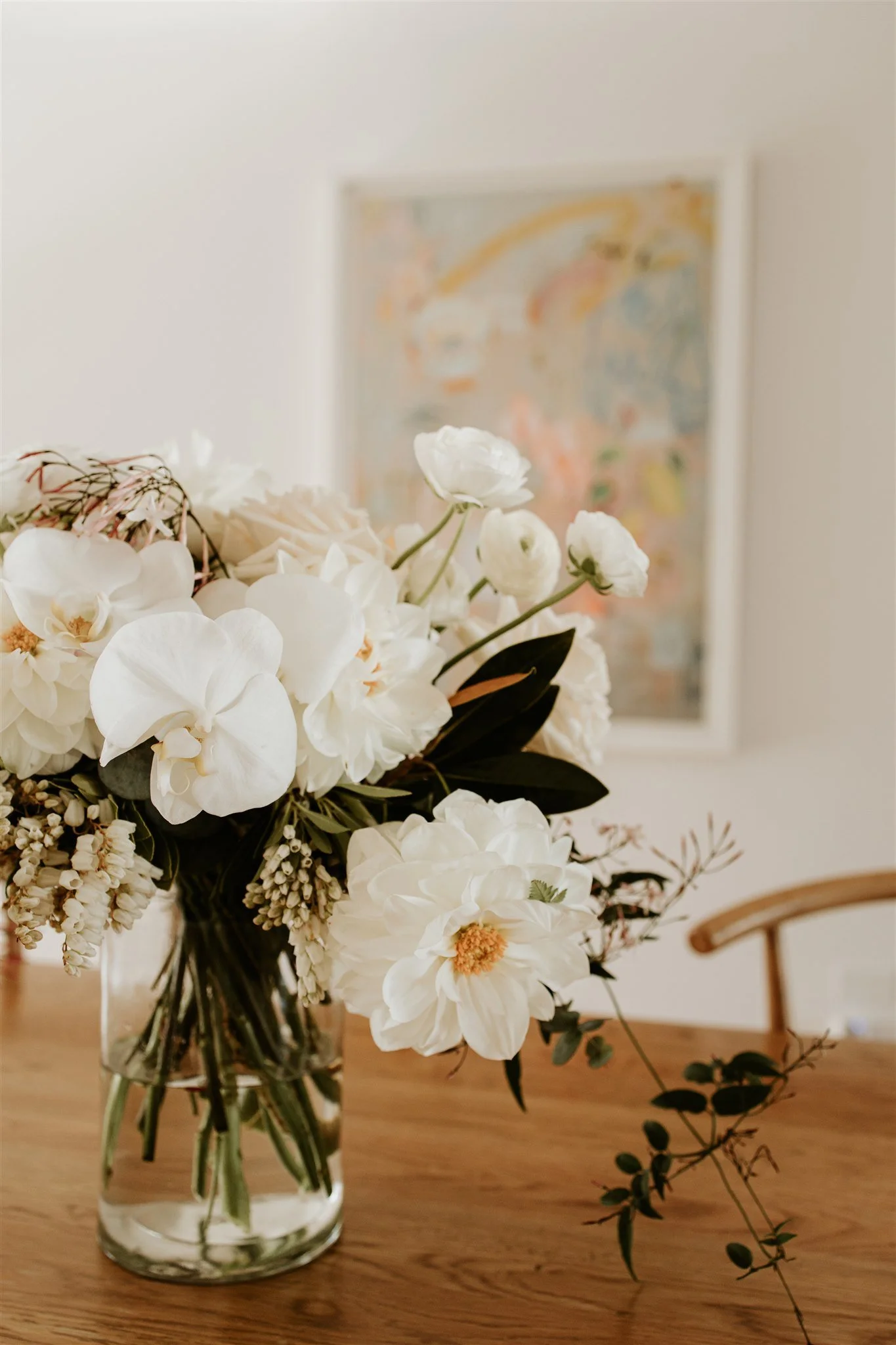 A clear glass vase filled with white flowers, including orchids, roses, and peonies, sitting on a wooden table with a blurred colorful abstract painting in the background.