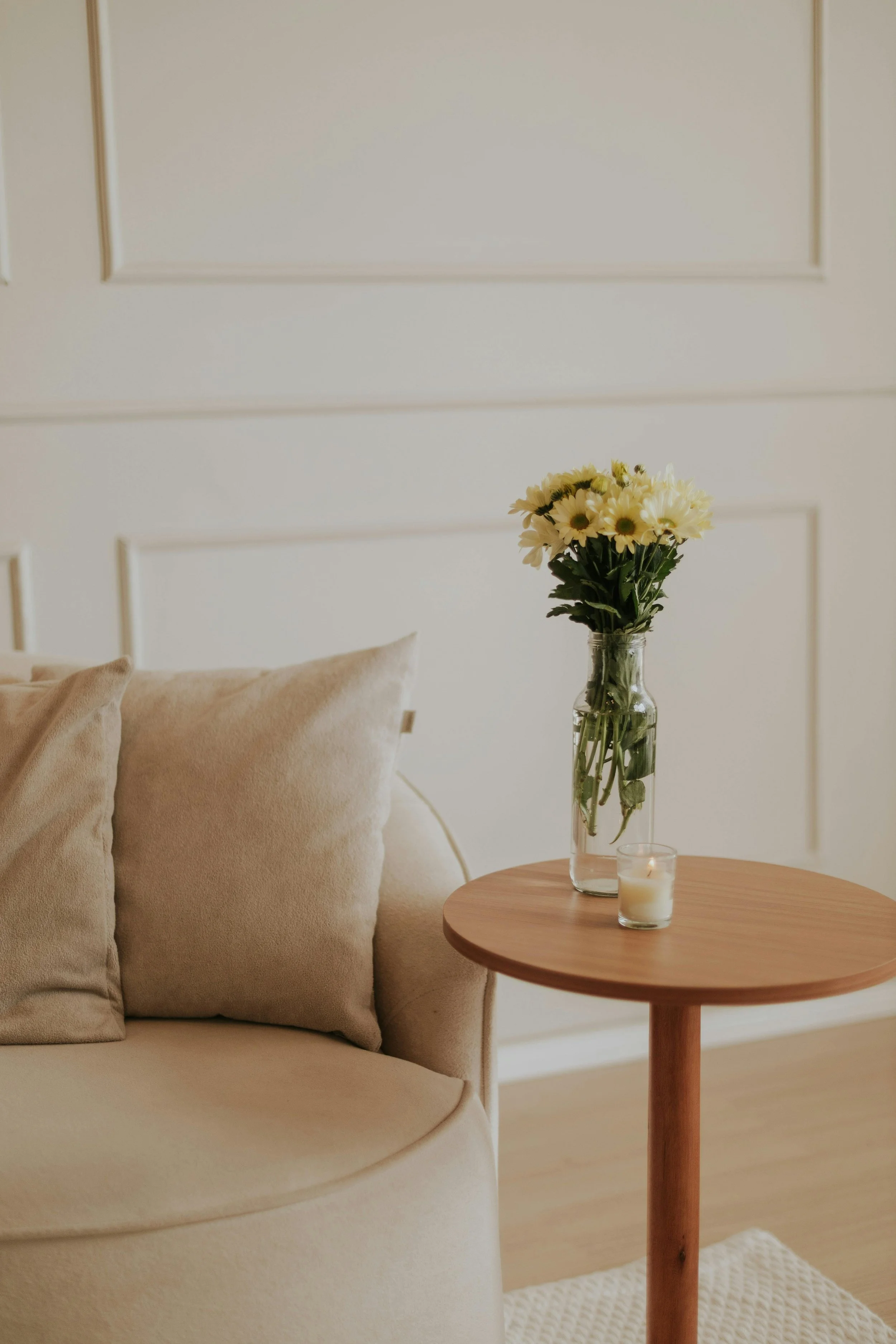 A beige sofa with matching cushions next to a small round wooden side table holding a glass vase with white and yellow flowers and a small candle in a glass holder, in a cozy living room.