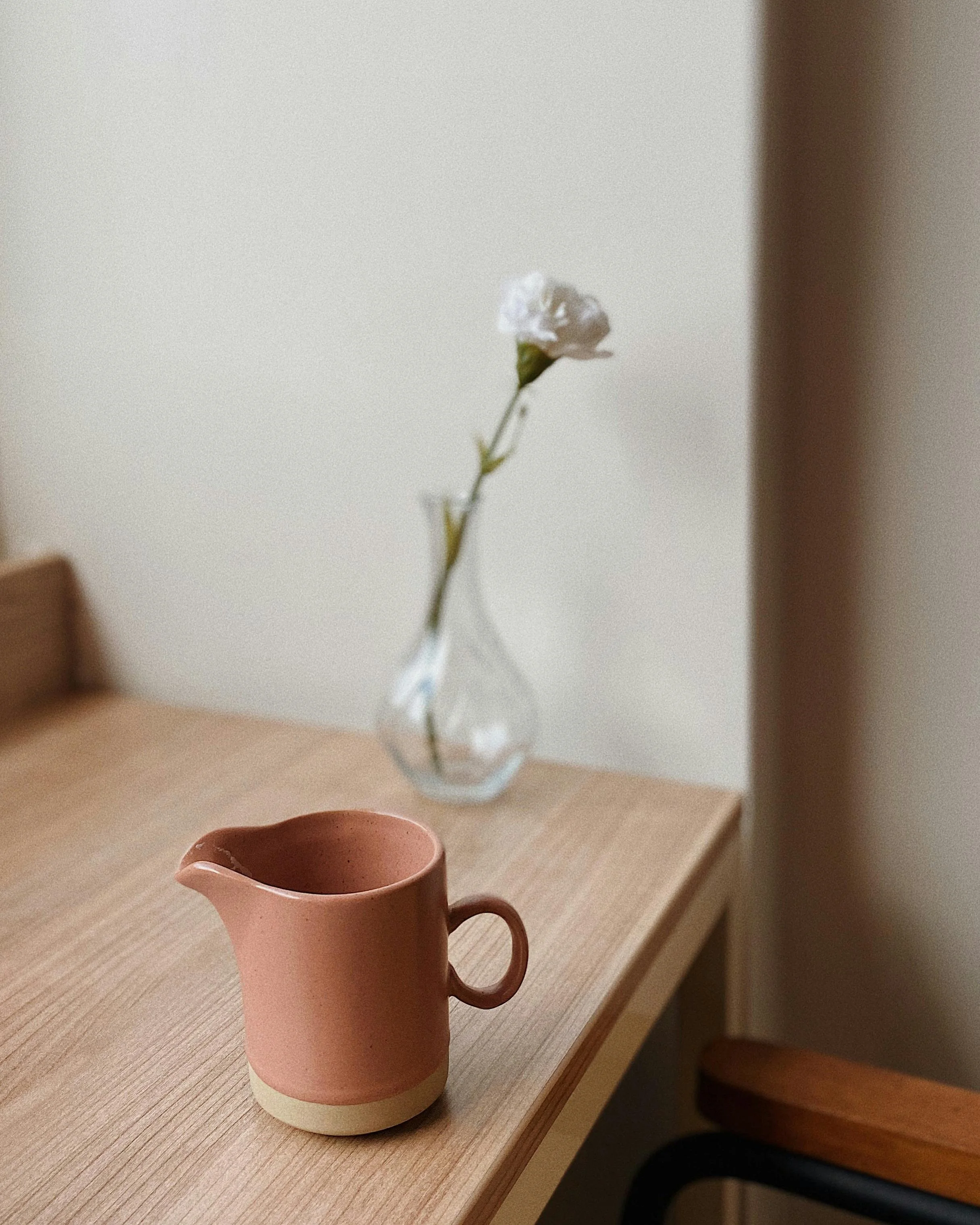 A pink ceramic pitcher with a beige base sitting on a wooden table, with a glass vase with a single white flower in the background.