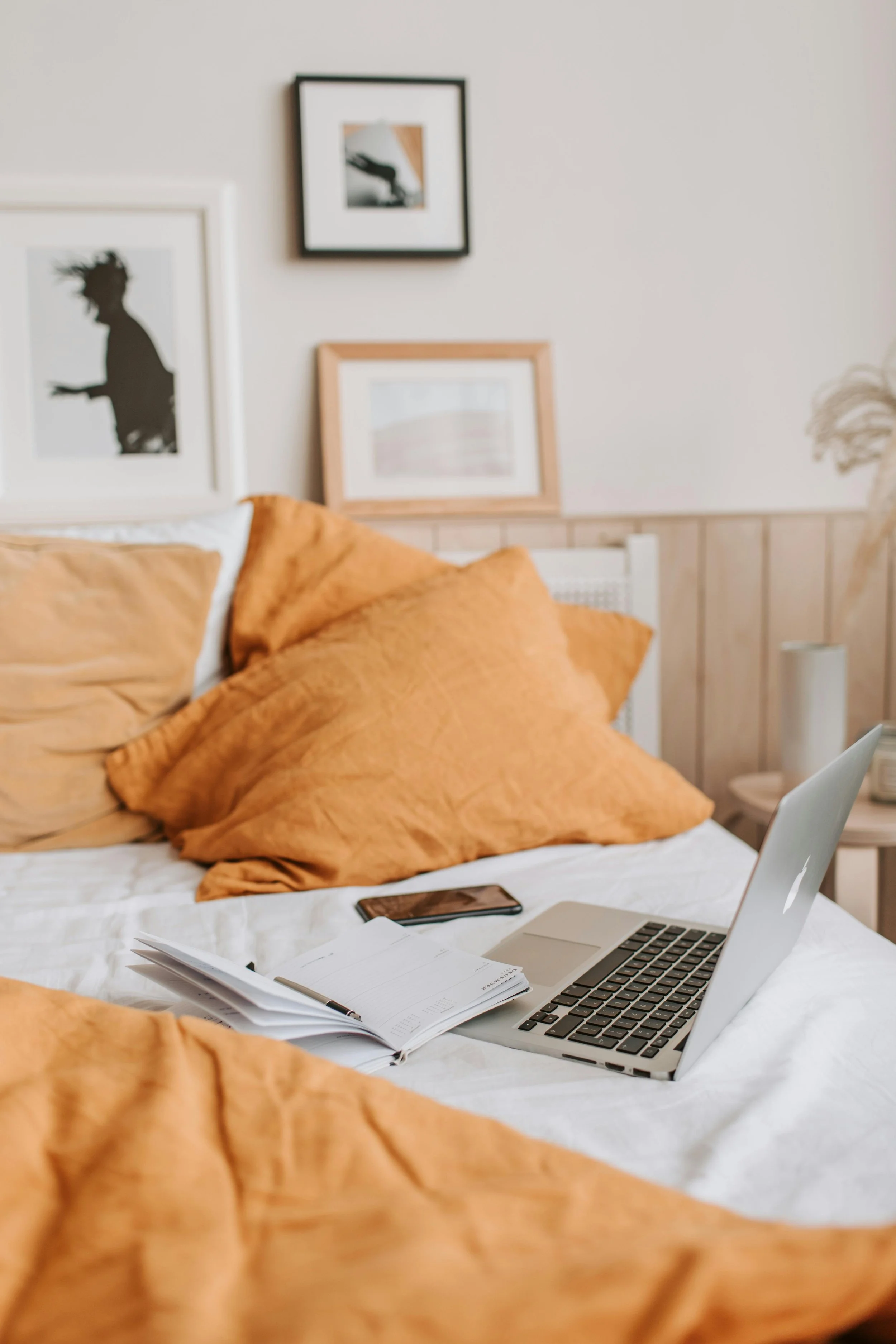 A neatly made bed with mustard-colored pillows, a laptop, open planner, and smartphone placed on top. The wall behind the bed features framed art, including a silhouette of a person with hair blowing, and other abstract pictures. There is a bedside table with a white lamp and a vase on the right.