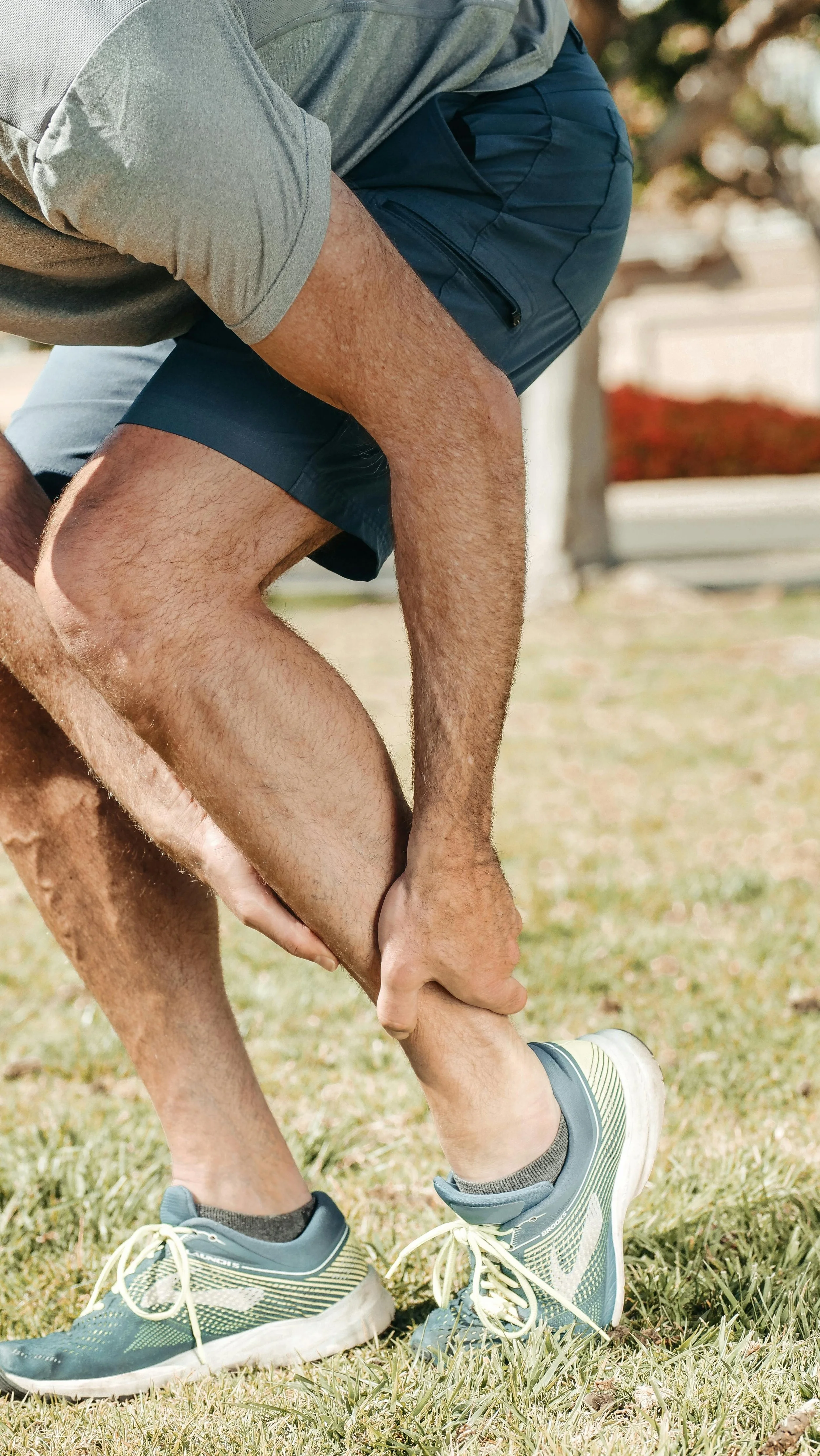 Close-up of a person tying their shoelace outdoors on a grassy area, wearing athletic shoes, shorts, and a gray shirt.