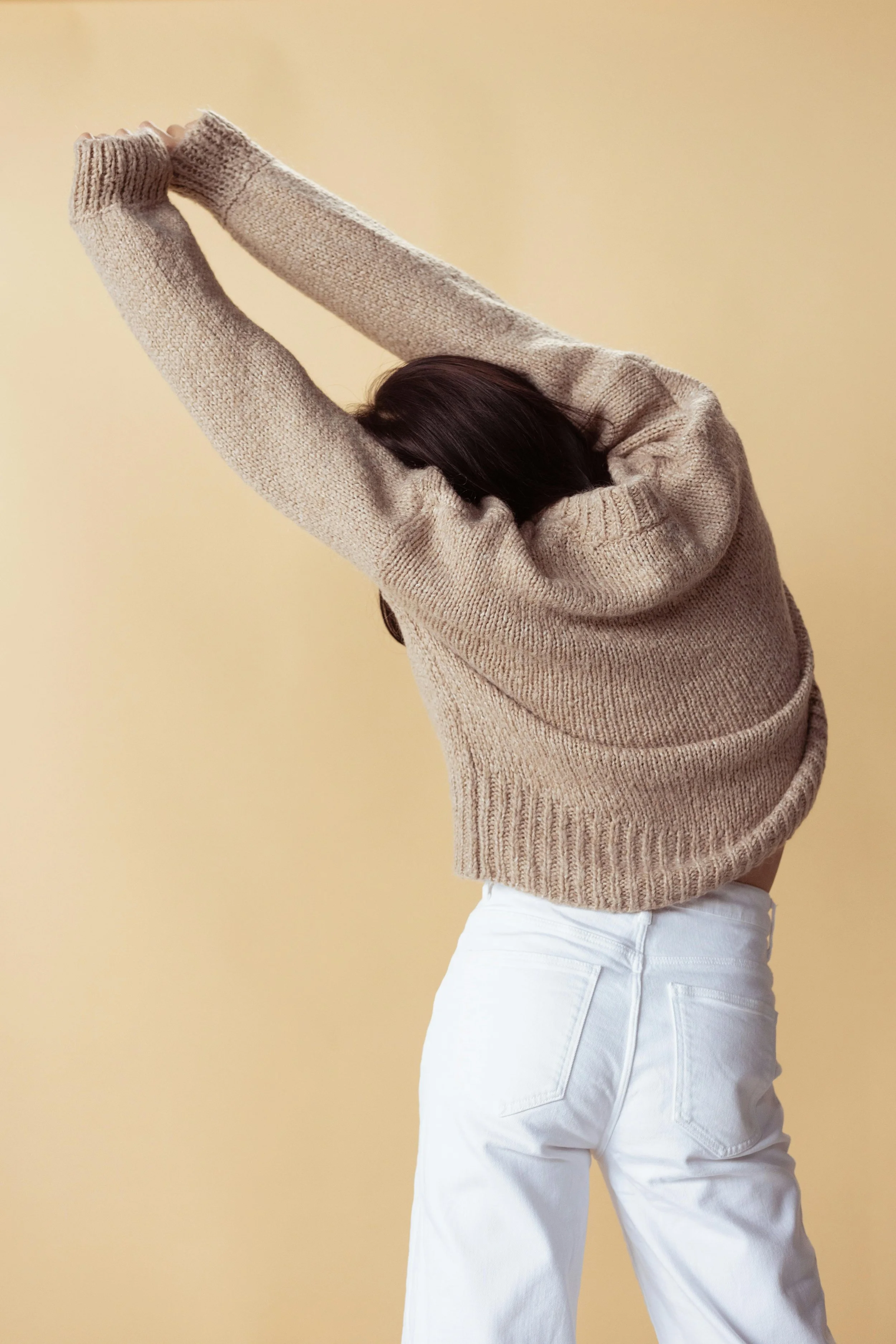 A woman stretching with her arms raised above her head, wearing a beige sweater and white pants, against a plain light yellow background.