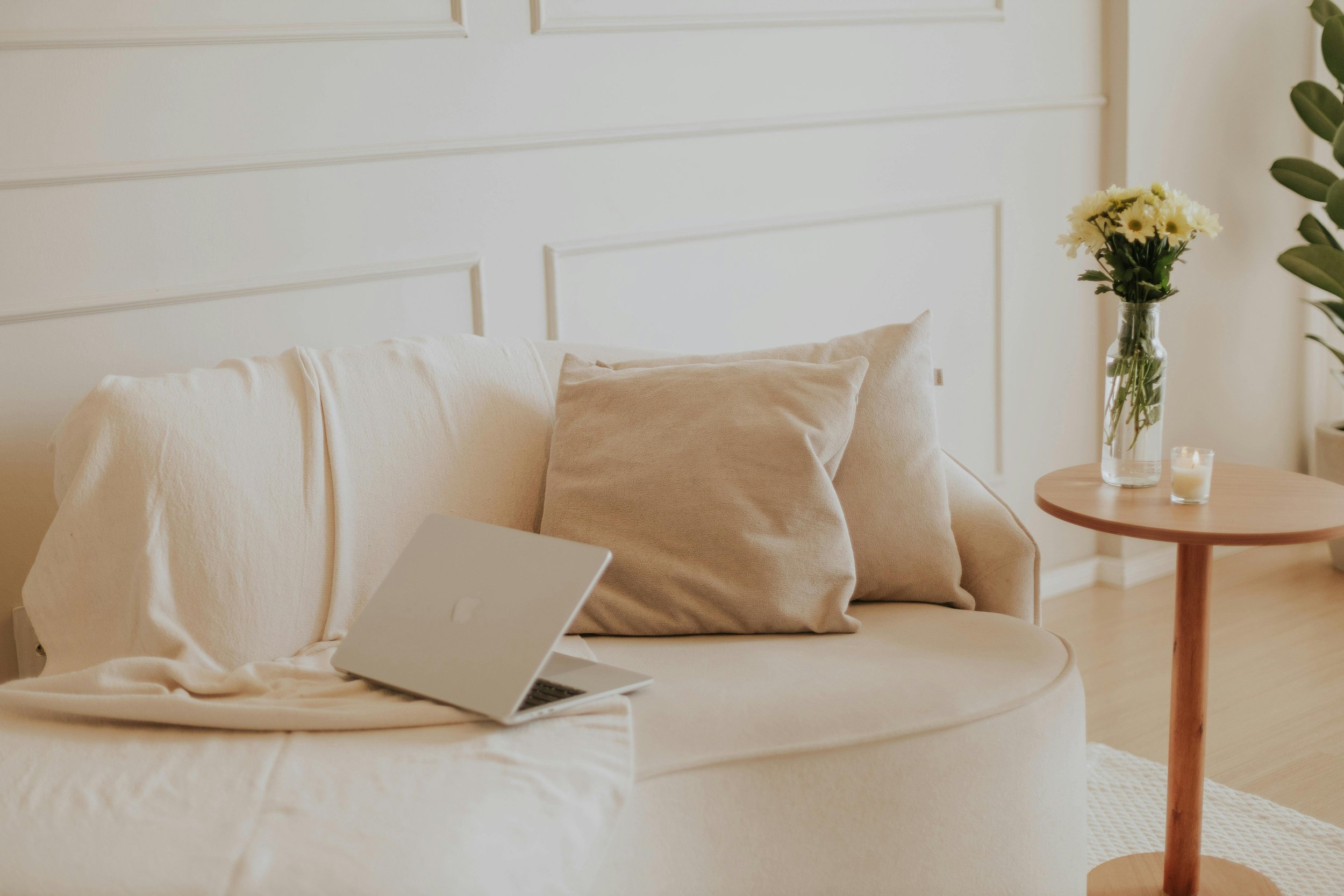 Cozy living room corner with a beige sofa, pillows, a laptop, a wooden side table with a vase of yellow flowers, a candle, and a plant in background.