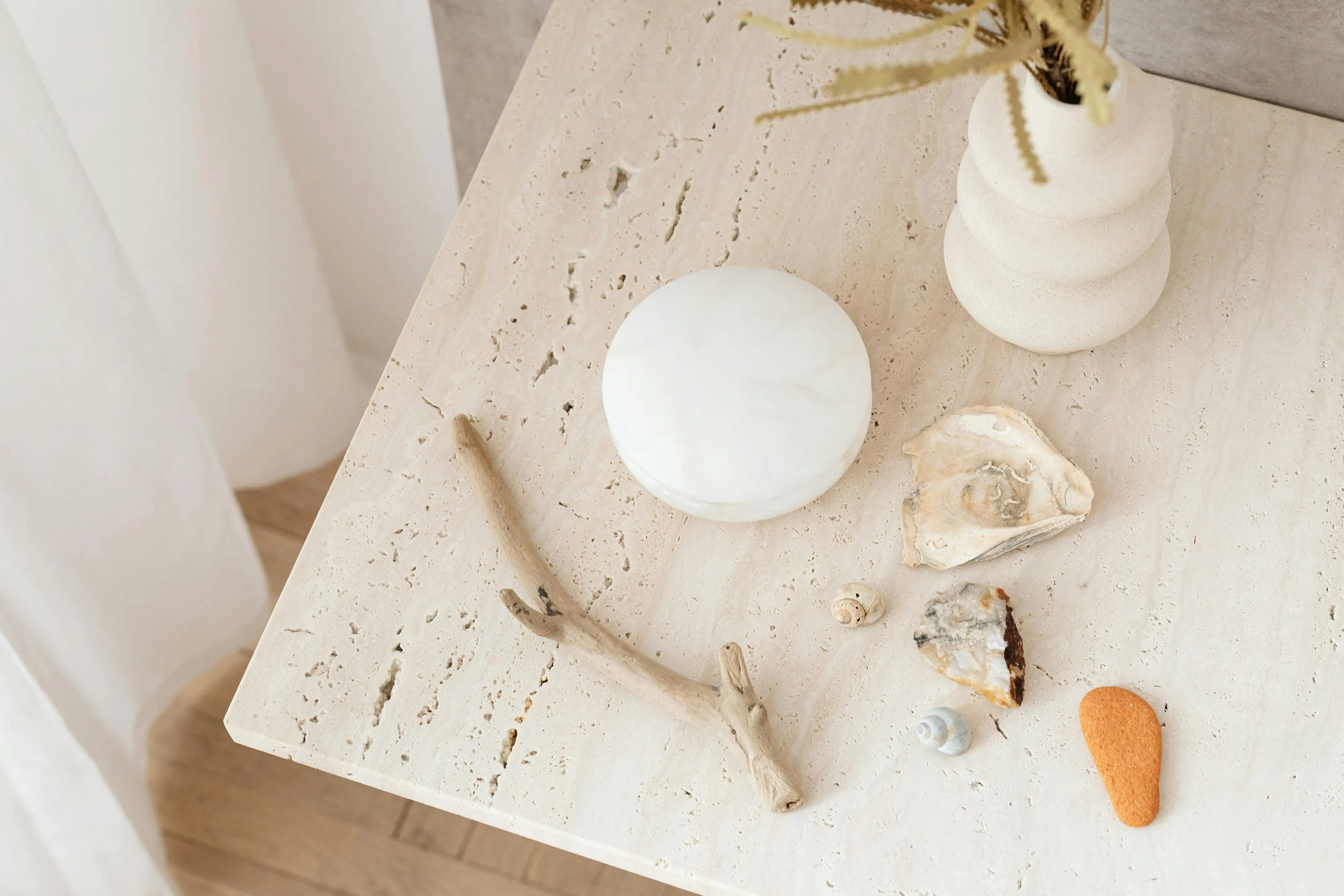 Decorative table arrangement with a white orb-shaped lamp, a textured white vase with dried plant, a piece of driftwood, a large shell, smaller shells, two stones, and a small orange object, on a light-colored textured surface.