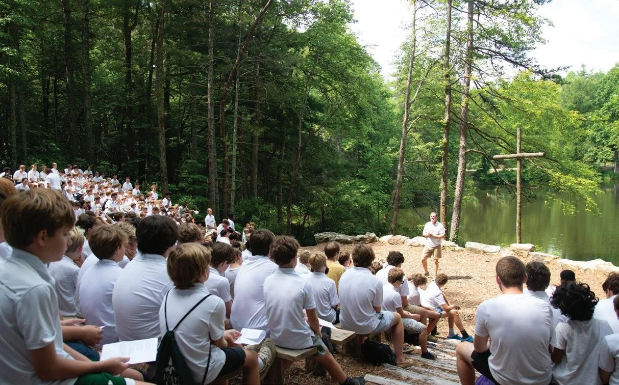 A large group of children and teenagers in white shirts sitting on benches outdoors near a lake, listening to a man speaking in front of a small cross and trees.
