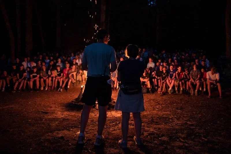 A large group of people sitting on benches around a campfire in a wooded outdoor setting at night, with two people standing and facing the fire and the crowd.