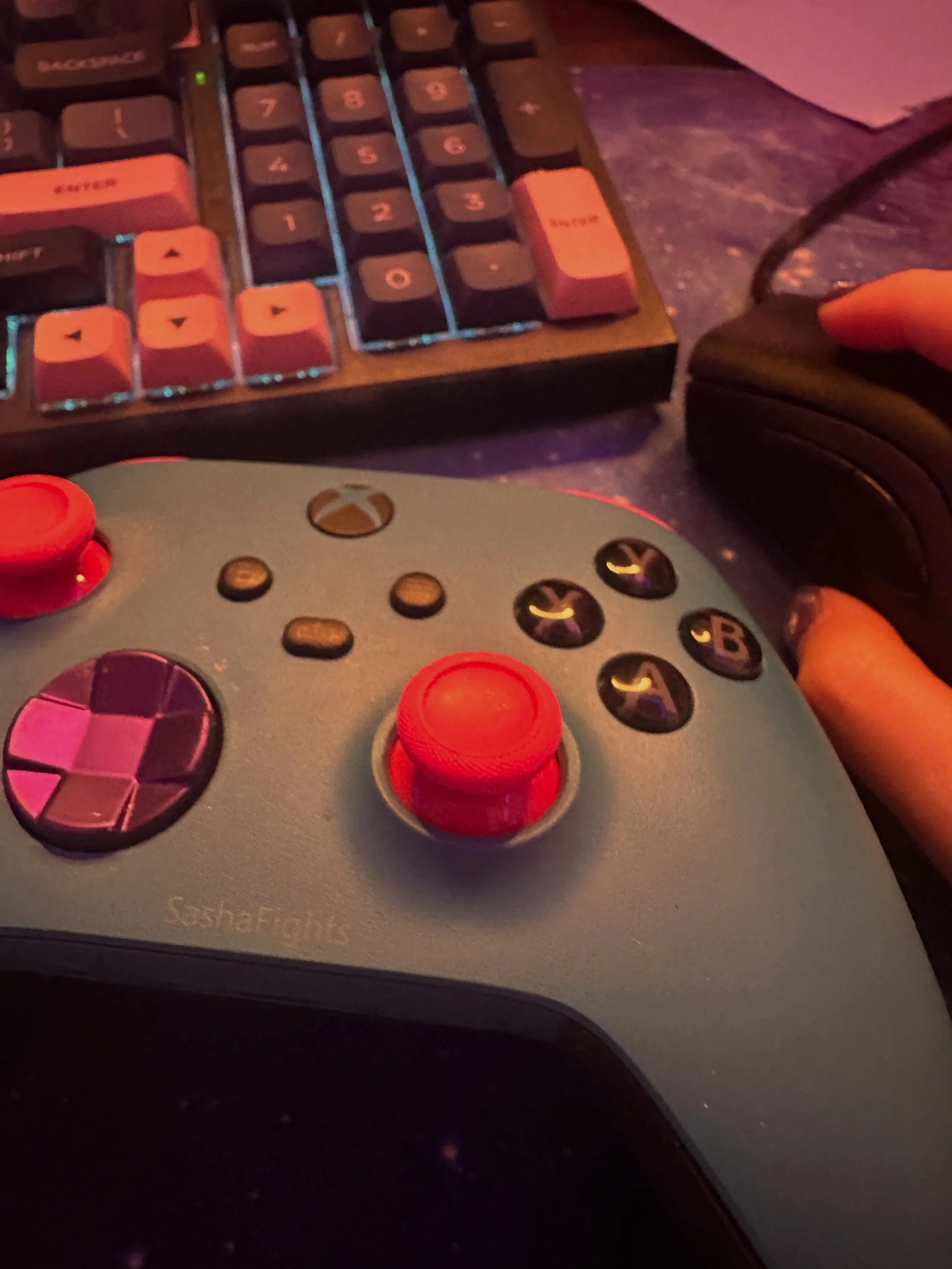 A gaming controller with red joysticks and purple buttons, a computer keyboard with pink keys, and a black computer mouse on a desk.