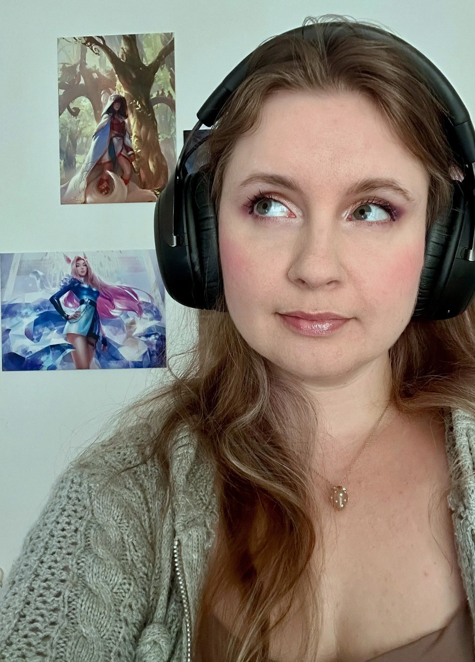 A woman wearing large black headphones with a beige knitted cardigan and a necklace with a leaf pendant, standing in front of a white wall with two fantasy-style posters.
