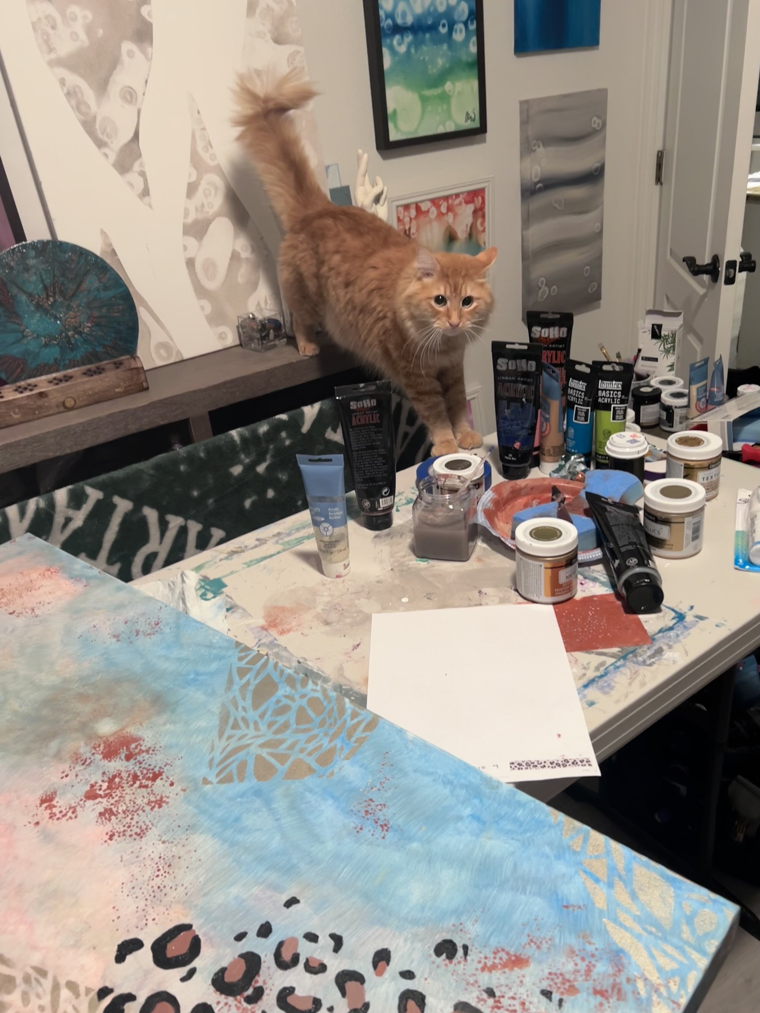 A fluffy orange cat standing on a cluttered table filled with paint supplies, in an art studio with colorful paintings on the wall.