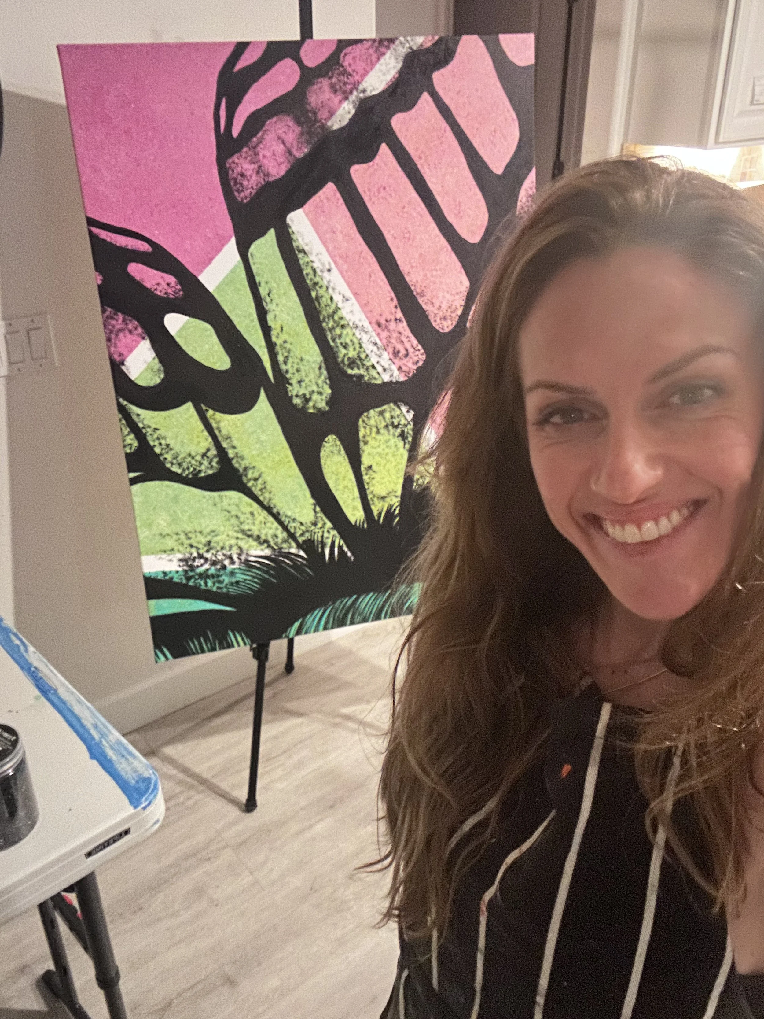 A woman smiling at a camera in front of a colorful abstract painting of a butterfly on an easel.