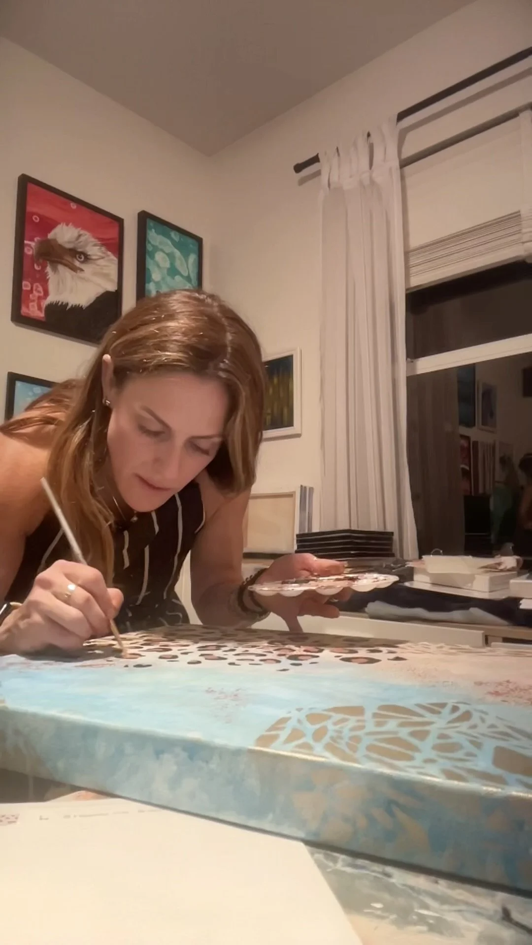 Woman painting or creating art on a large canvas or surface at a table in a room with wall art, including an eagle's head painting.