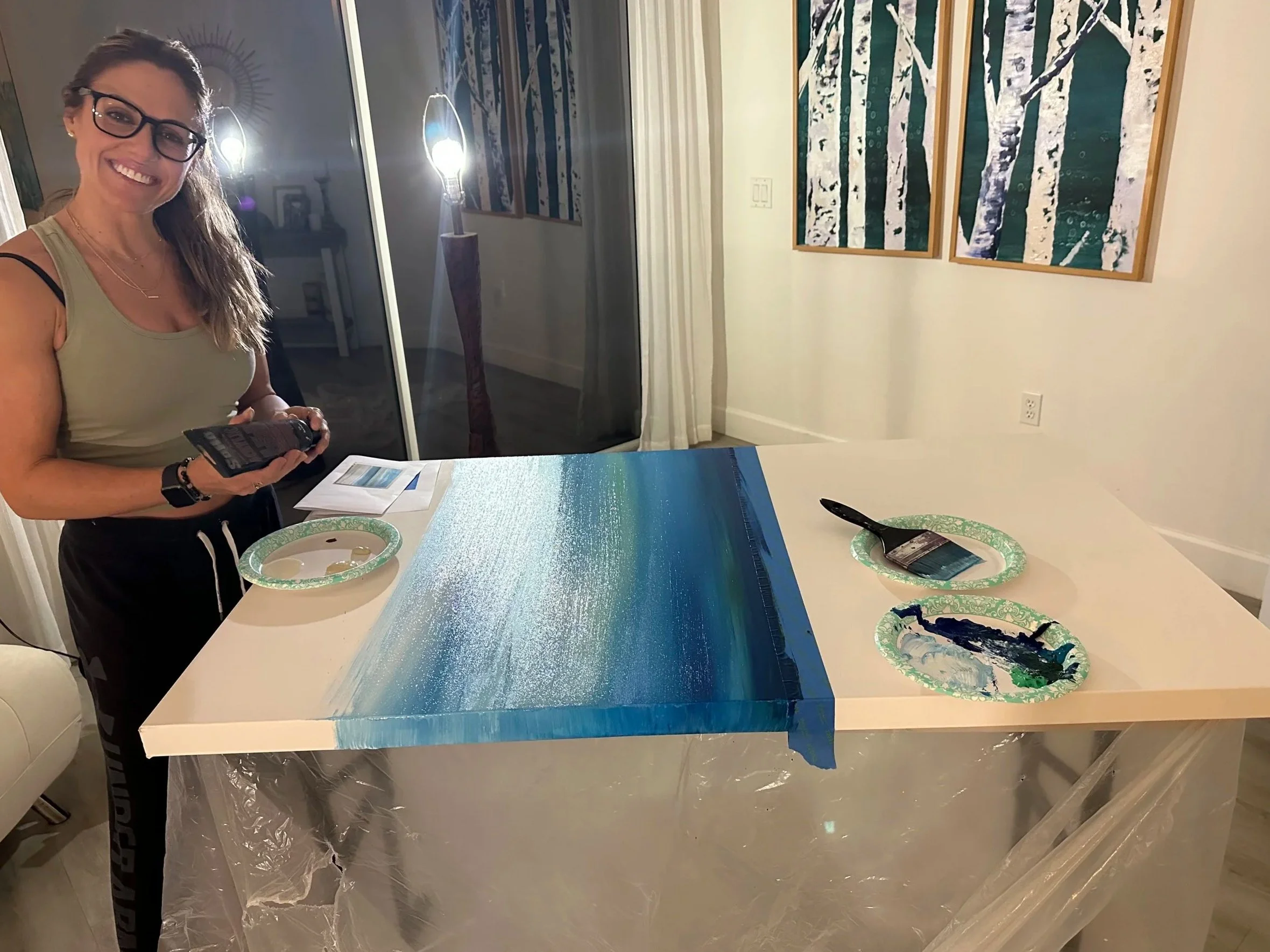 Woman standing at a table with a partially painted landscape canvas, holding a paint tube, with art supplies and paintings of trees on the wall behind her.