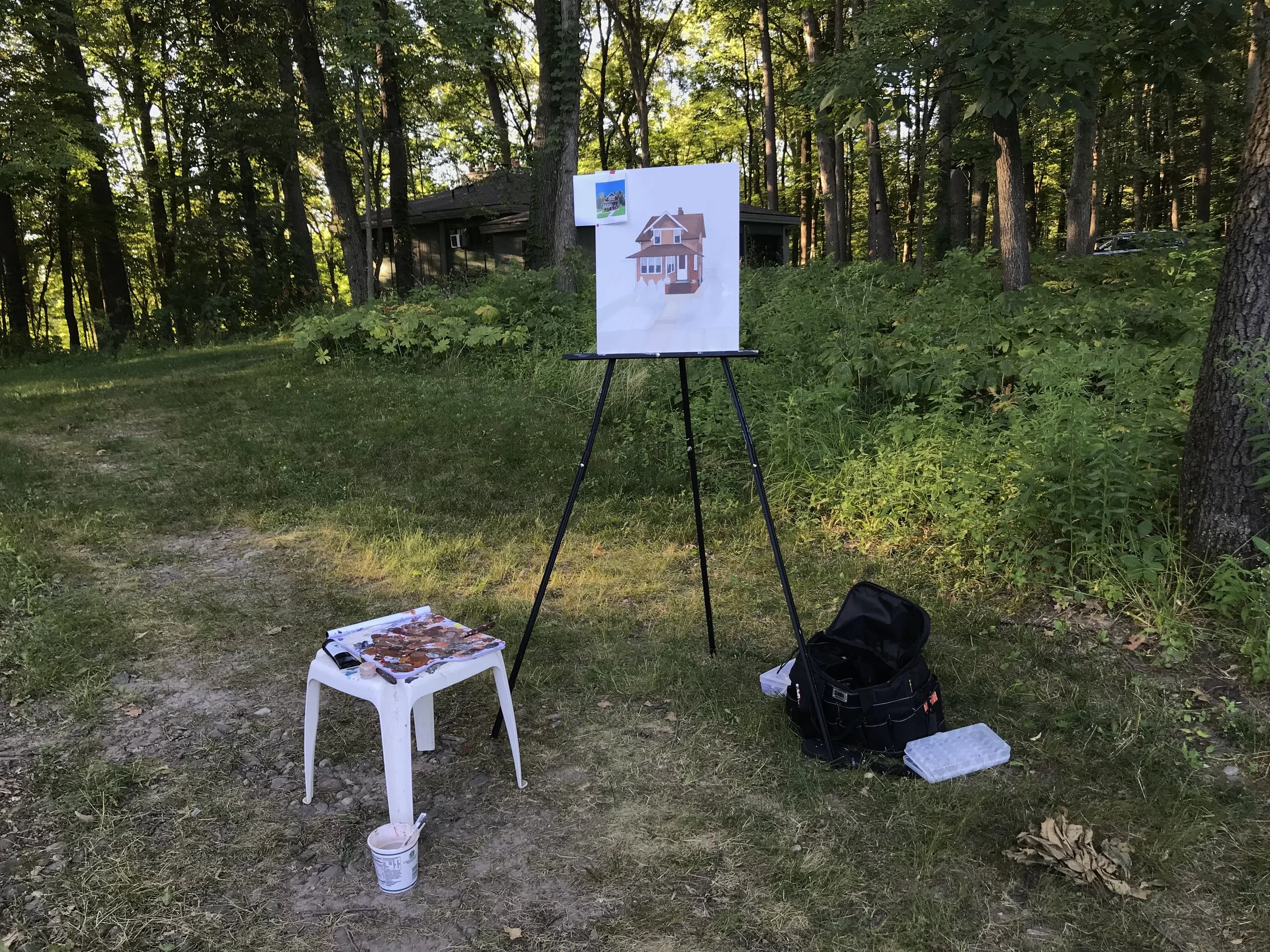 An outdoor scene with a painting easel displaying a painting of a house, a small white stool with paint supplies, a black bag, and a plastic container, set in a wooded area with trees and greenery.