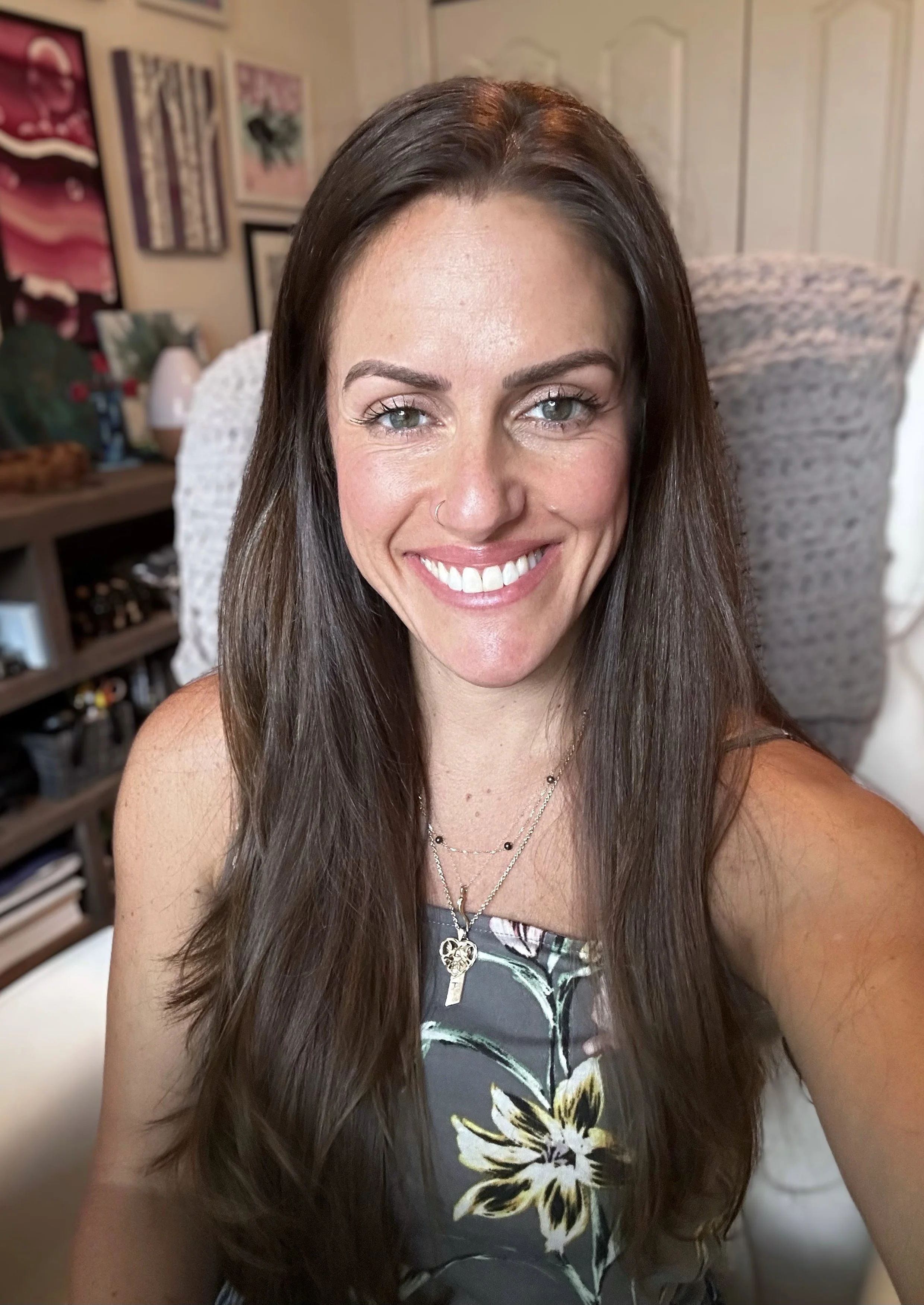 A woman with long dark hair, light complexion, and blue eyes smiling at the camera in a room with artwork on the walls and a bookshelf in the background.