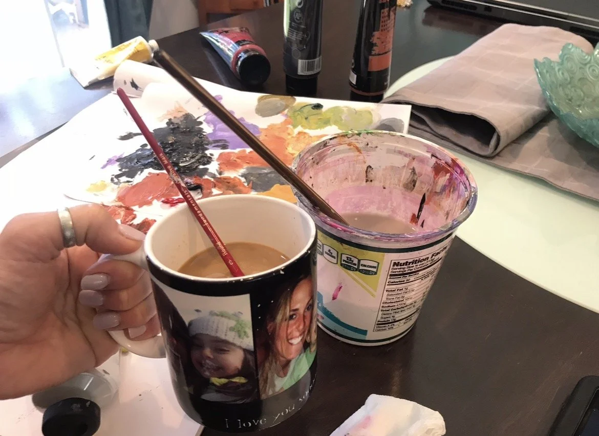 A person holding a mug filled with coffee, and a paintbrush accidentally put into it. The mug is on a work area with a paint palette, brushes, and paint containers. There are paint tubes and a napkin nearby.