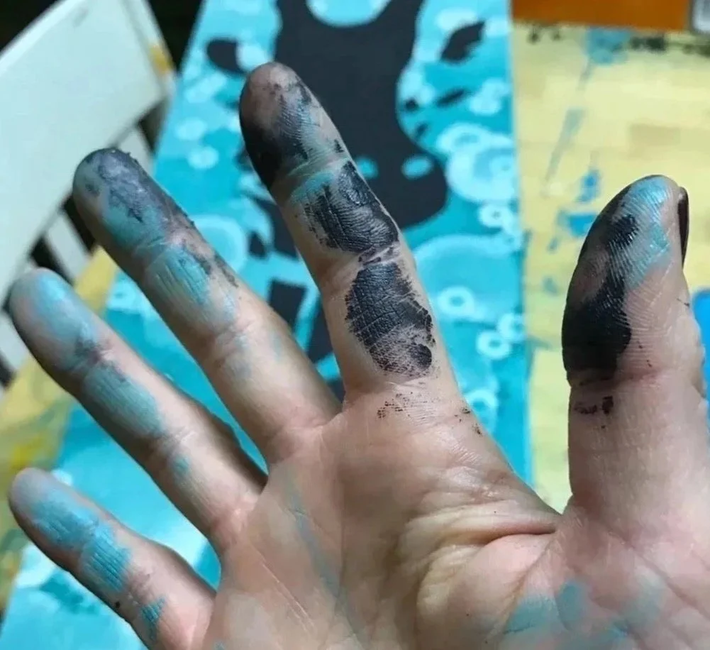 A person's hand covered in black and blue paint stains, with a colorful, patterned canvas in the background.