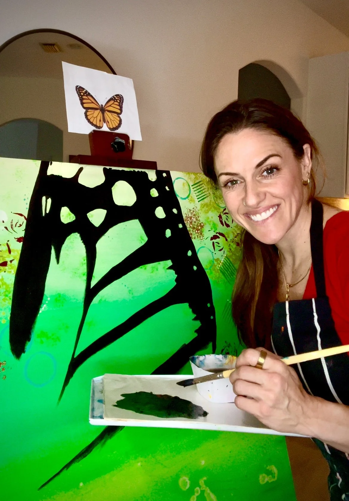 A woman painting a large butterfly on a vibrant green abstract canvas, with a small reference image of a butterfly on top of her easel.