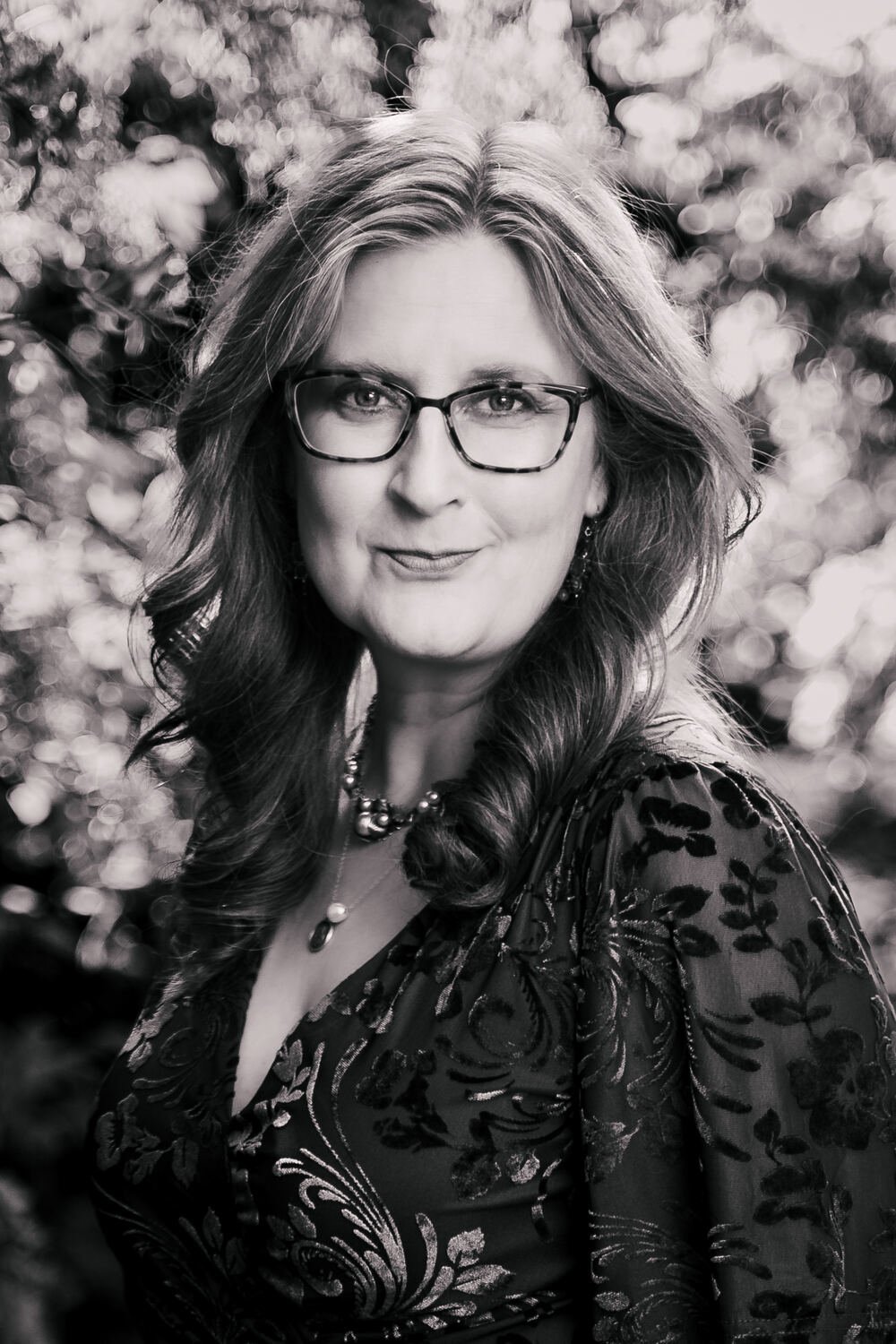 A black and white portrait of a woman with glasses, wavy shoulder-length hair, wearing a patterned dress and jewelry, with blurred foliage in the background.
