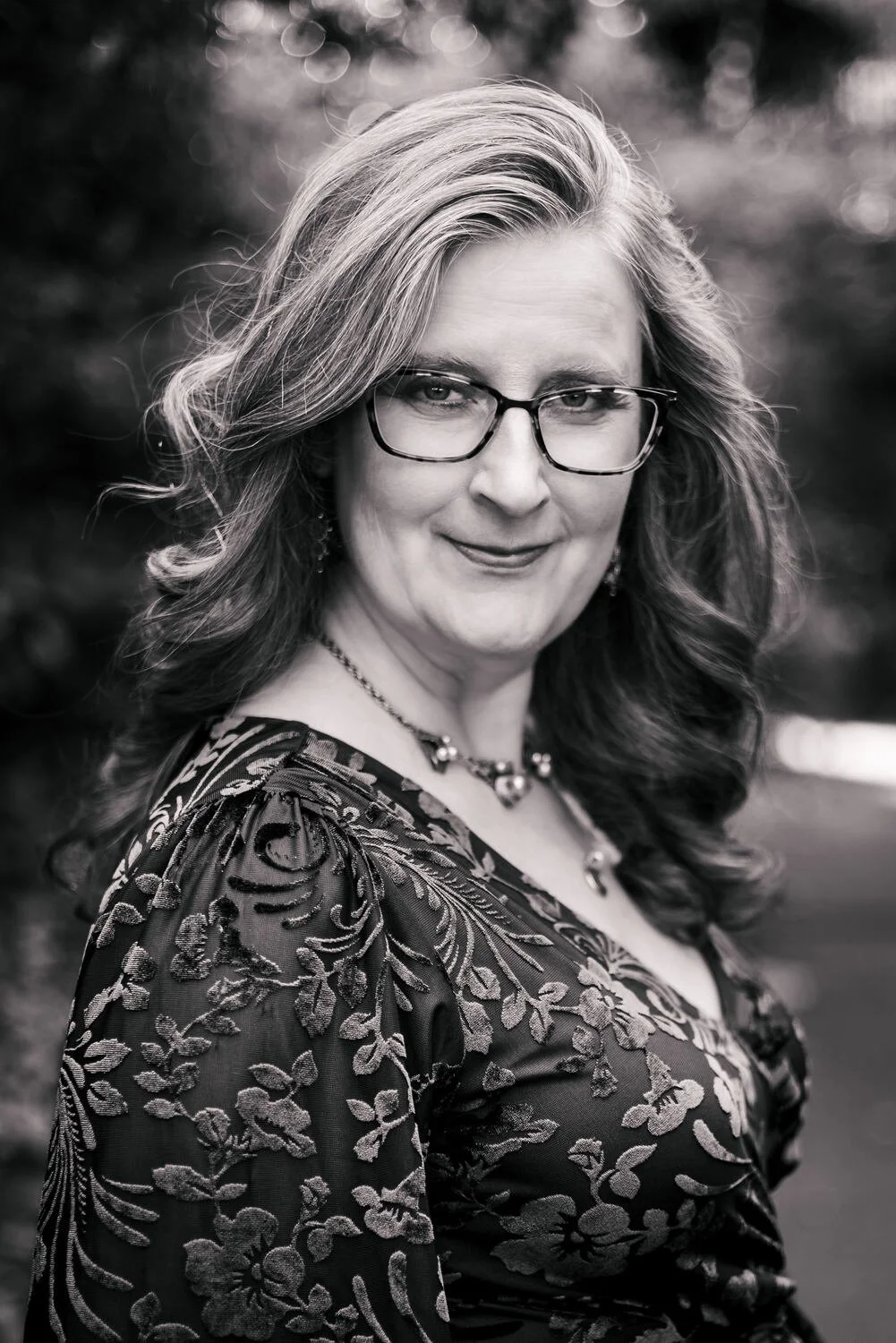 Black and white portrait of a woman with wavy hair, wearing glasses, a patterned top, and jewelry, outdoors with blurred background.
