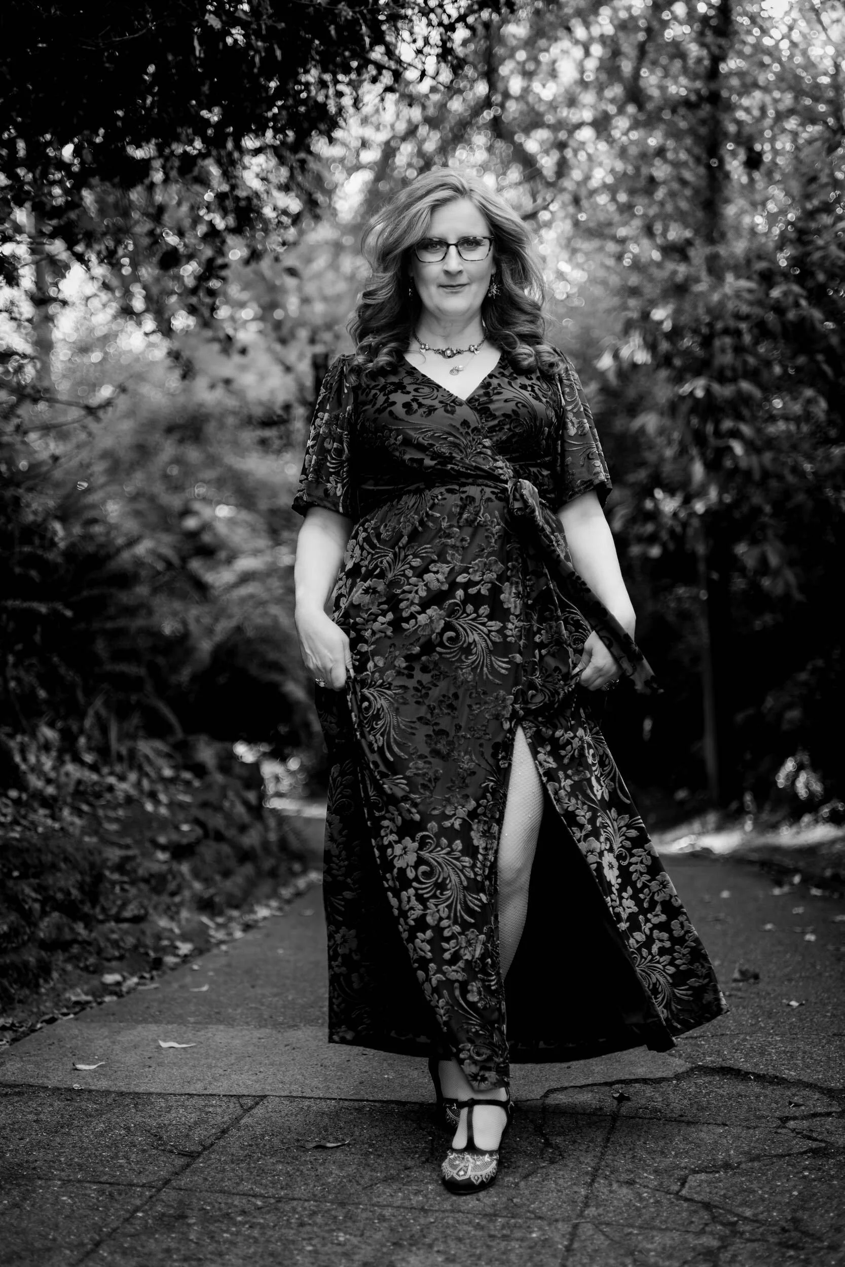 A woman walking on a paved path in an outdoor setting, dressed in a long, floral patterned dress with a slit, wearing high heels, sunglasses, and jewelry, surrounded by trees and foliage.