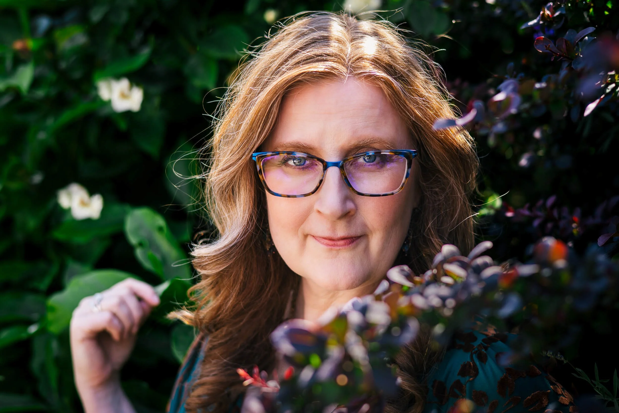 A woman with long, wavy red hair and glasses, standing outdoors among green and purple foliage, looking at the camera.
