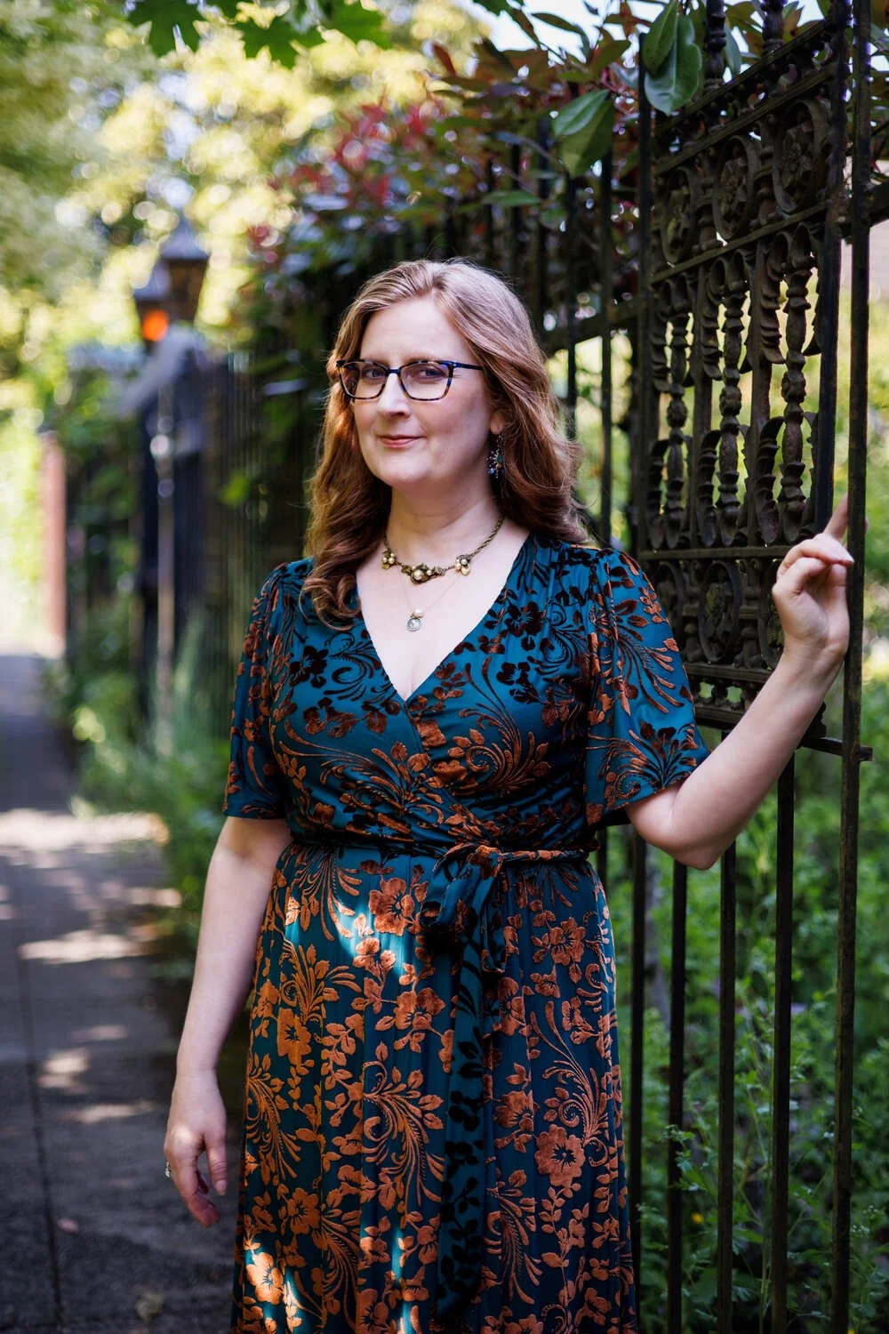 A woman standing outside next to a black wrought-iron fence, wearing glasses and a blue dress with brown floral patterns, with trees and greenery in the background.