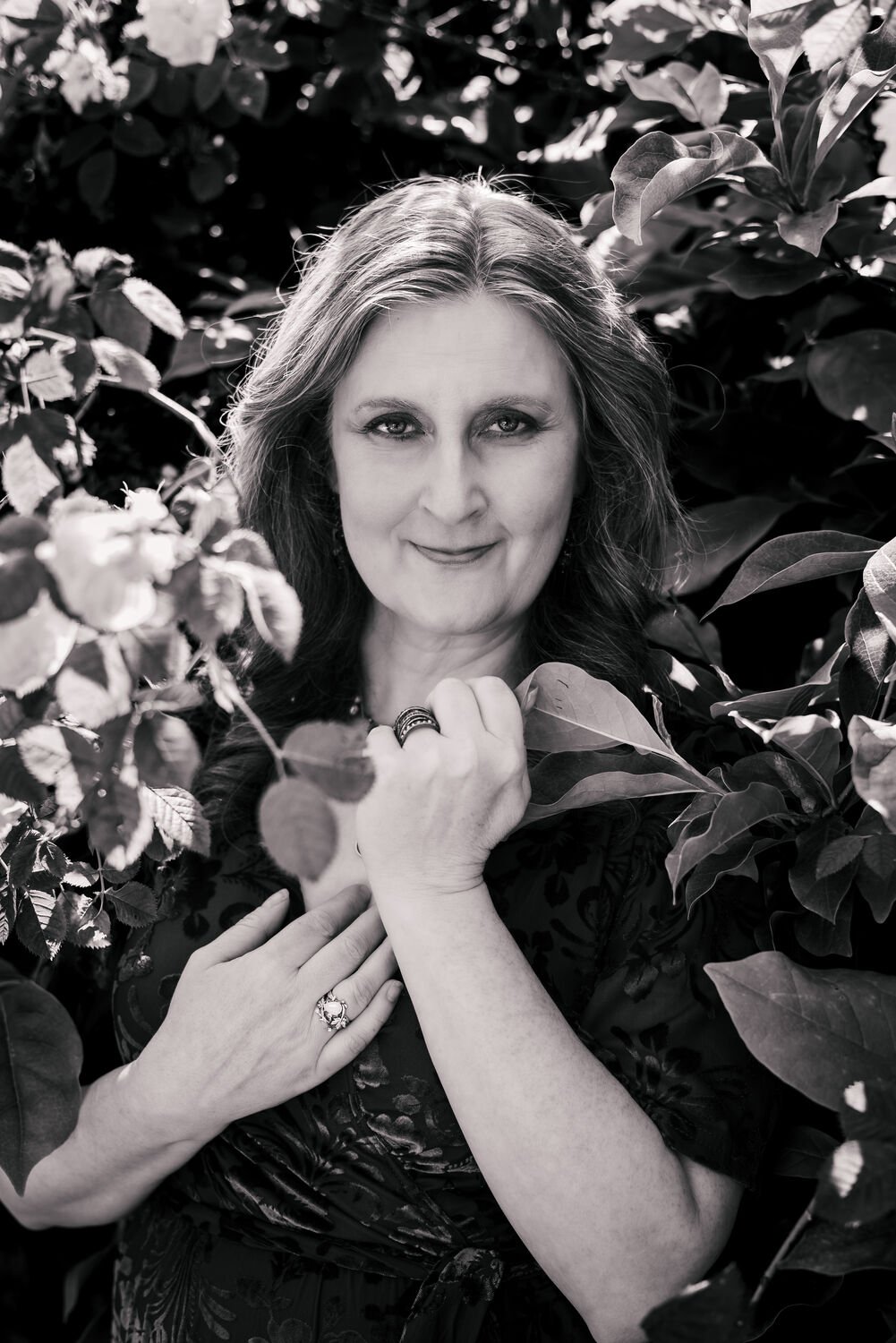 Black and white photo of a woman with long hair, smiling, surrounded by leaves and foliage, with her hands gently touching her chest and face.