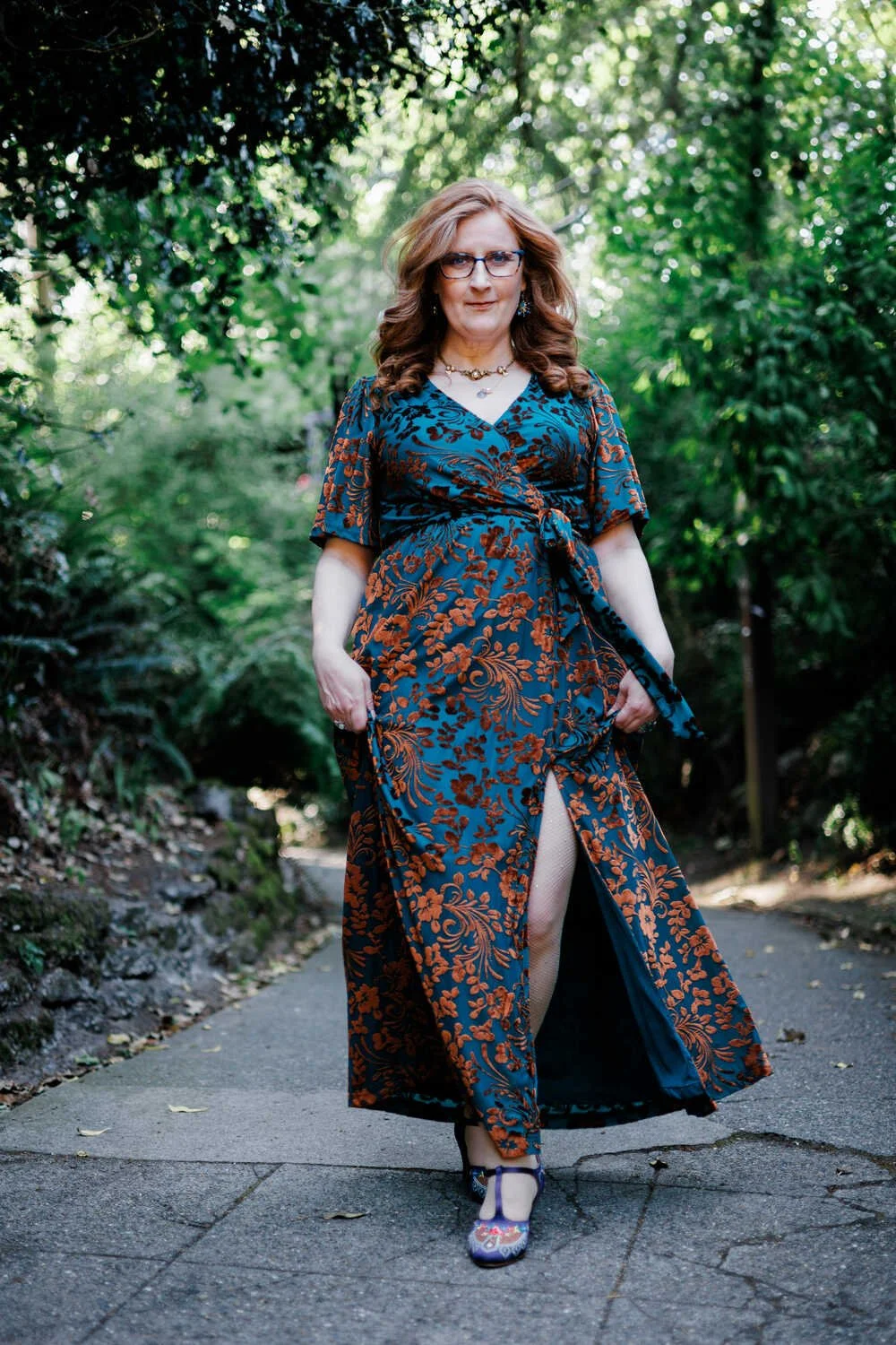 A woman with curly red hair, glasses, and a necklace, wearing a long blue dress with an orange floral pattern, walking on a paved path surrounded by green foliage.