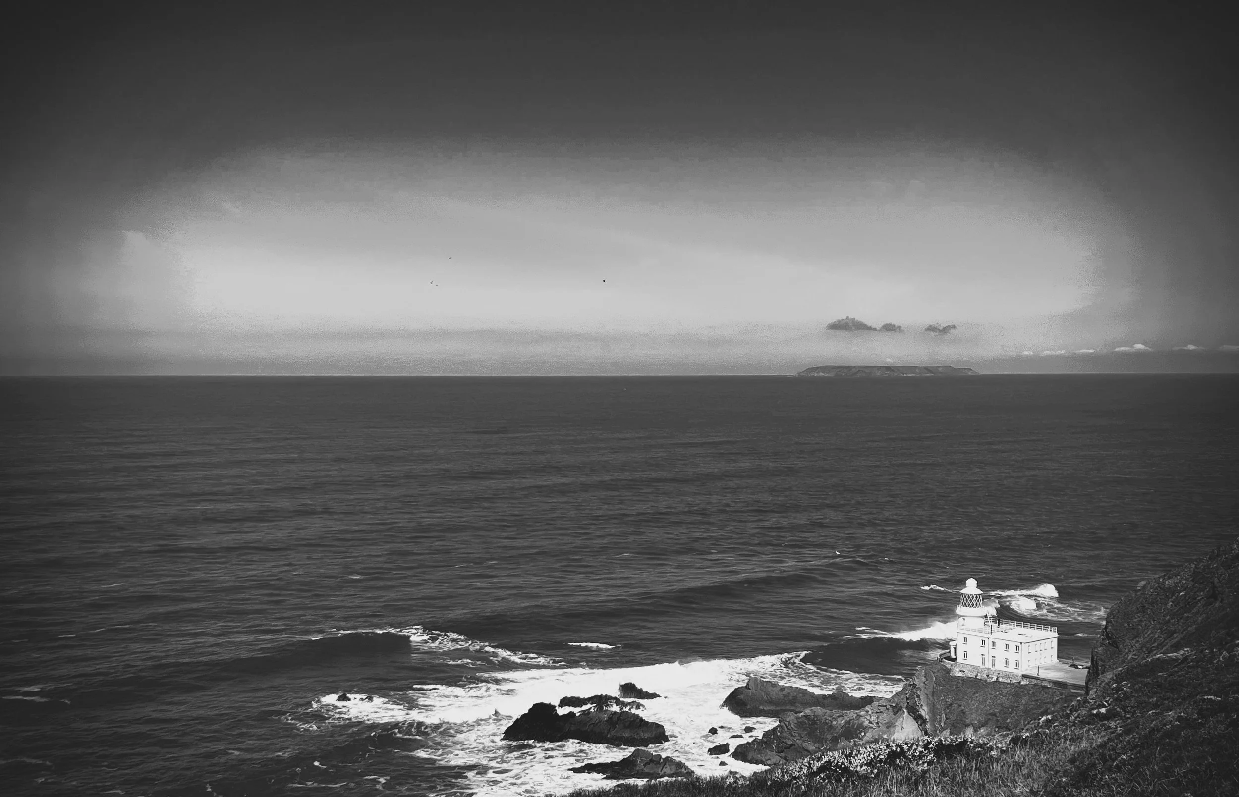 Black and white photo of a Hartland Lighthouse (credit:  Mark Burnett) on a rocky coastline (North Devon, UK) with the ocean and distant island (Lundy Island) in the background.