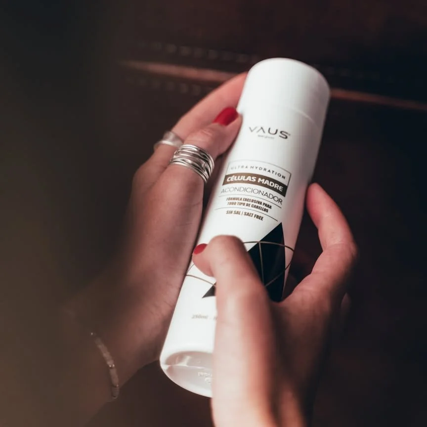 Mãos segurando um spray de condicionador VAUS, com foco na embalagem branca e rótulo preto e dourado, com fundo escuro
