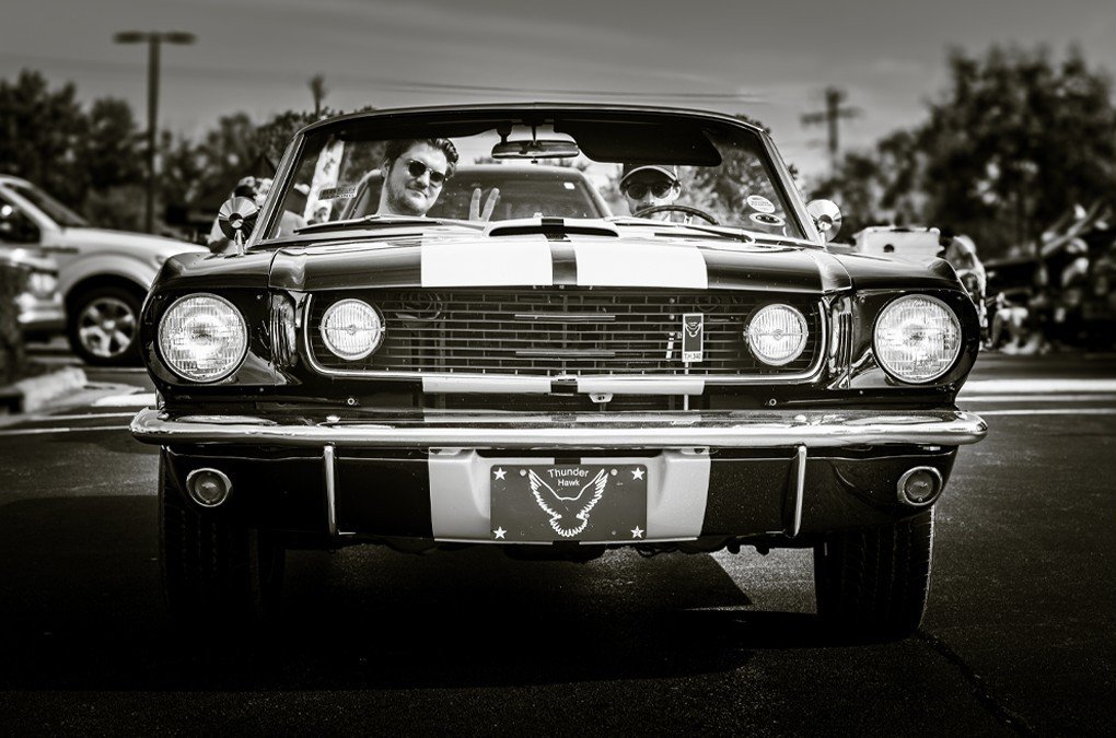 Black and white photo of two men sitting in a vintage car with a racing stripe design on the hood. The car is parked in a lot, with other vehicles and trees in the background. The license plate features an eagle and the words "Thunder Hawk."