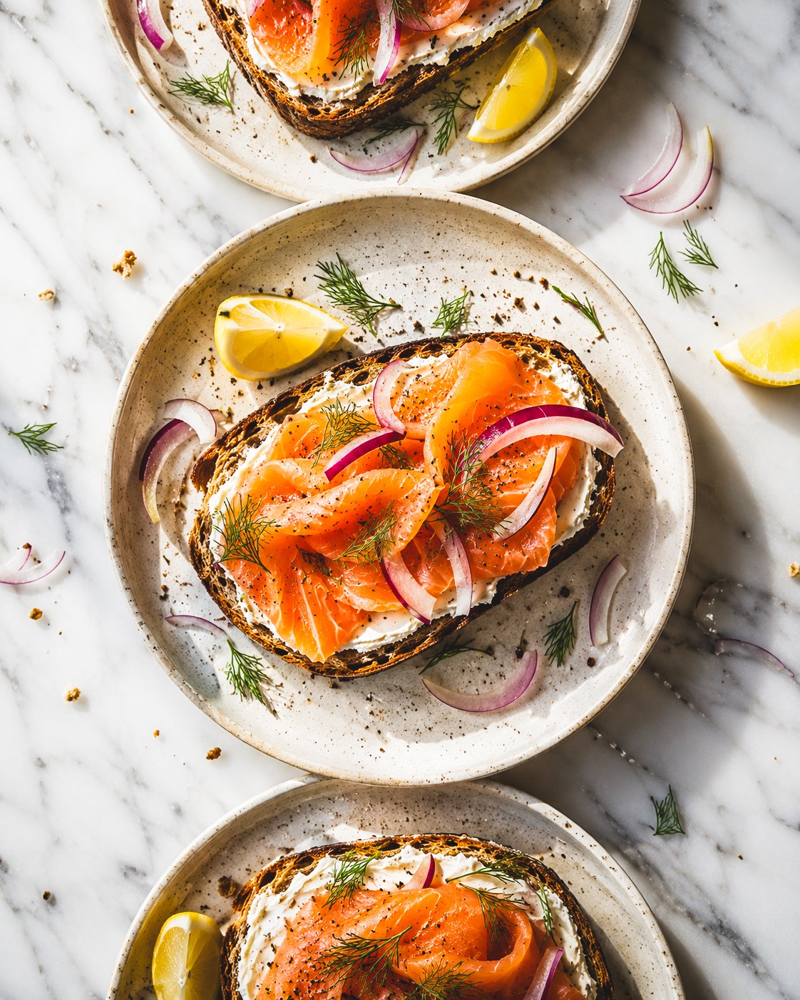 Three plates of bagels with cream cheese, smoked salmon, red onion slices, dill, and lemon wedges on a marble surface.
