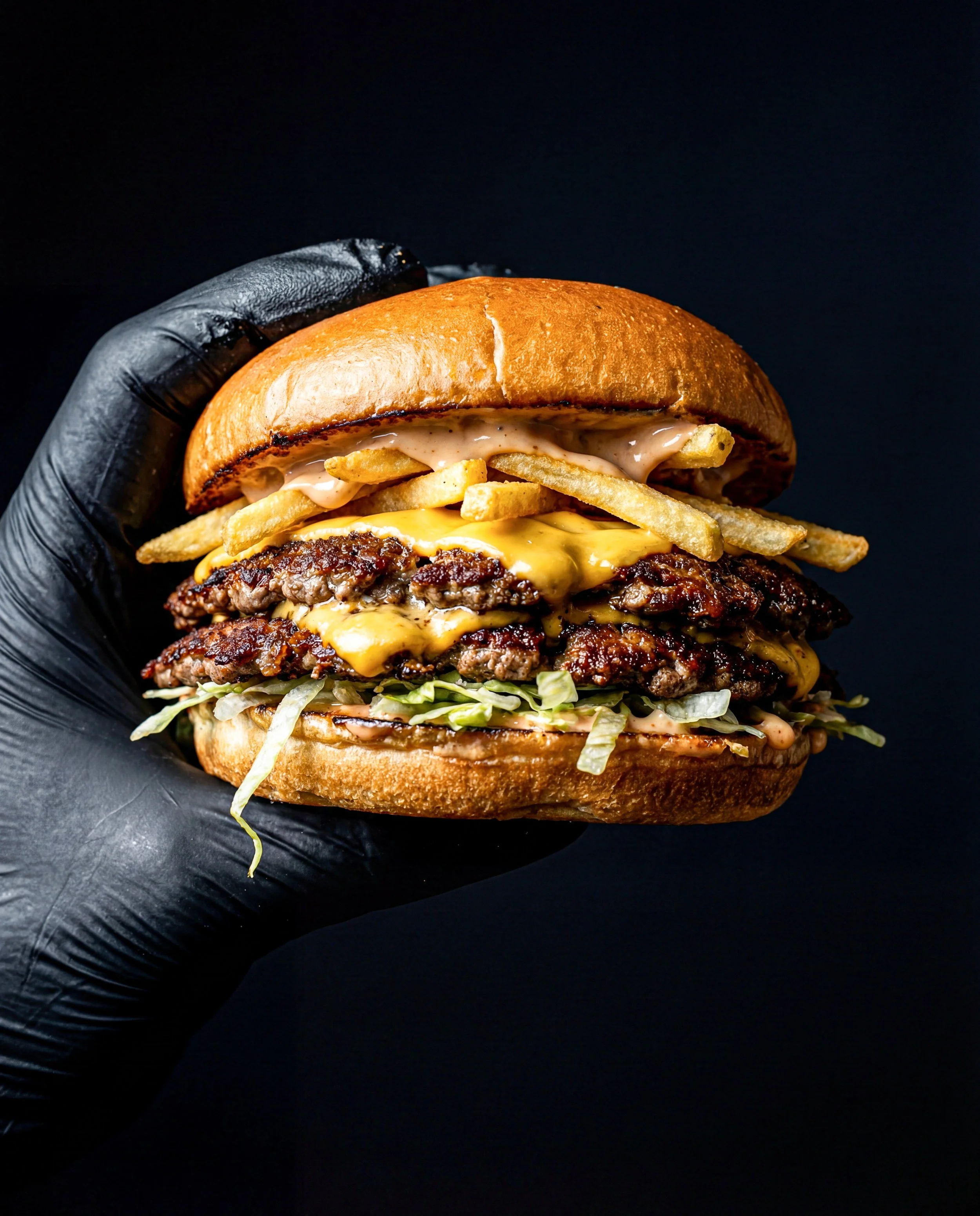 Close-up of a loaded cheeseburger with multiple beef patties, melted cheese, French fries, shredded lettuce, creamy sauce, all inside a toasted bun, against a black background.